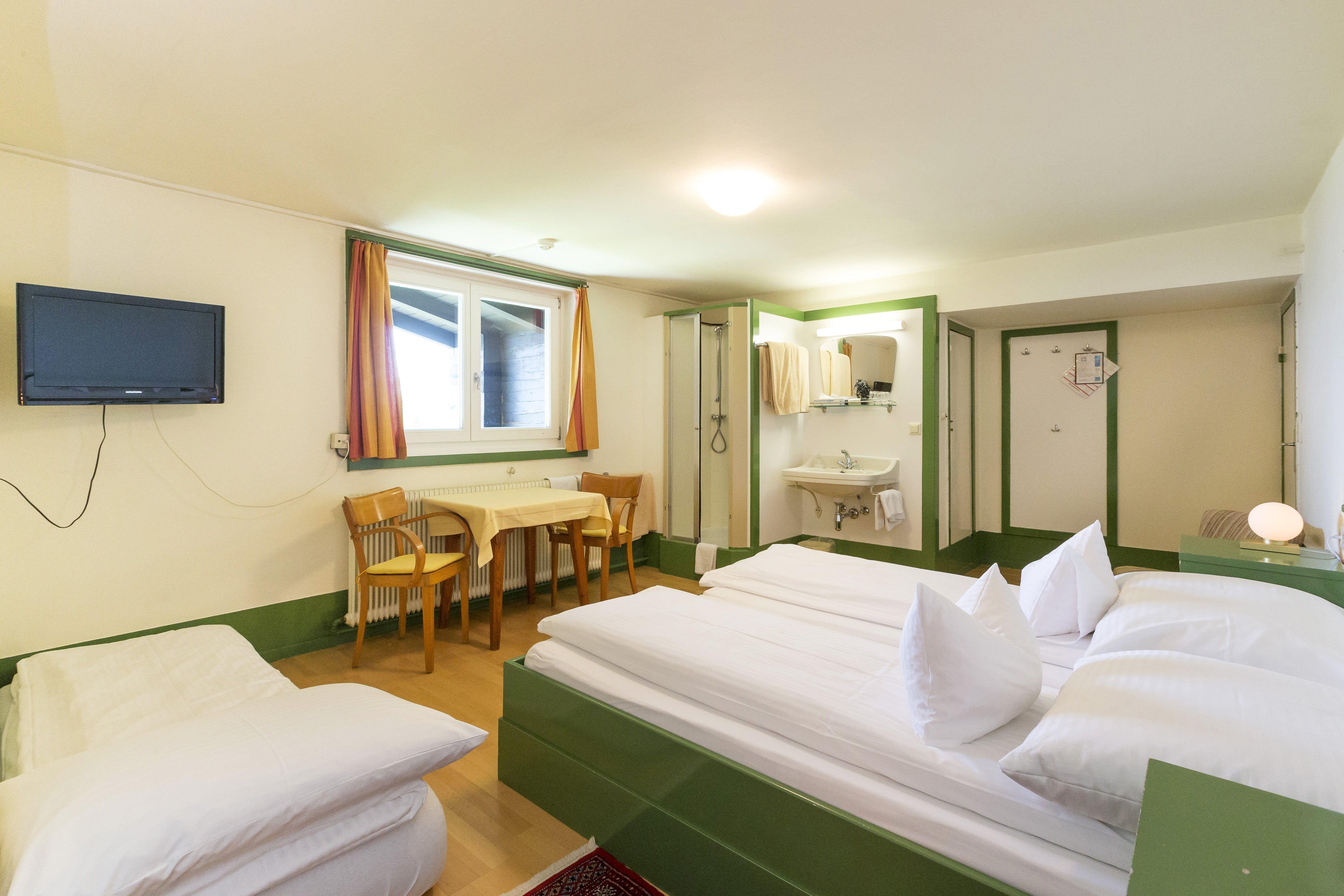 A simply furnished hotel room with a double bed, table, chairs, TV and washing area.
