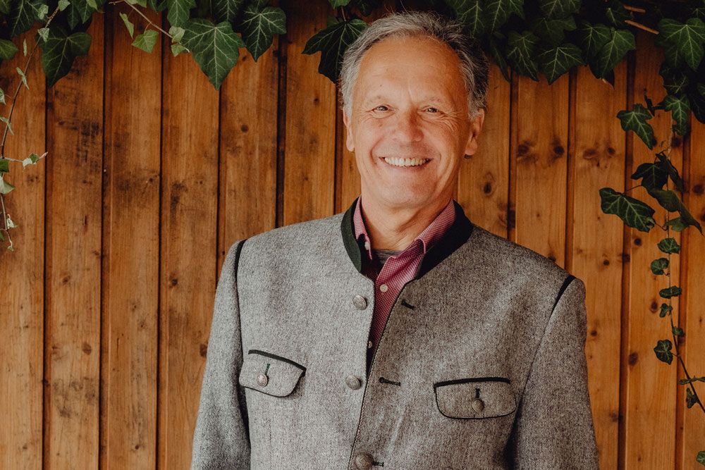 A smiling man in traditional dress in front of a wooden wall with ivy.