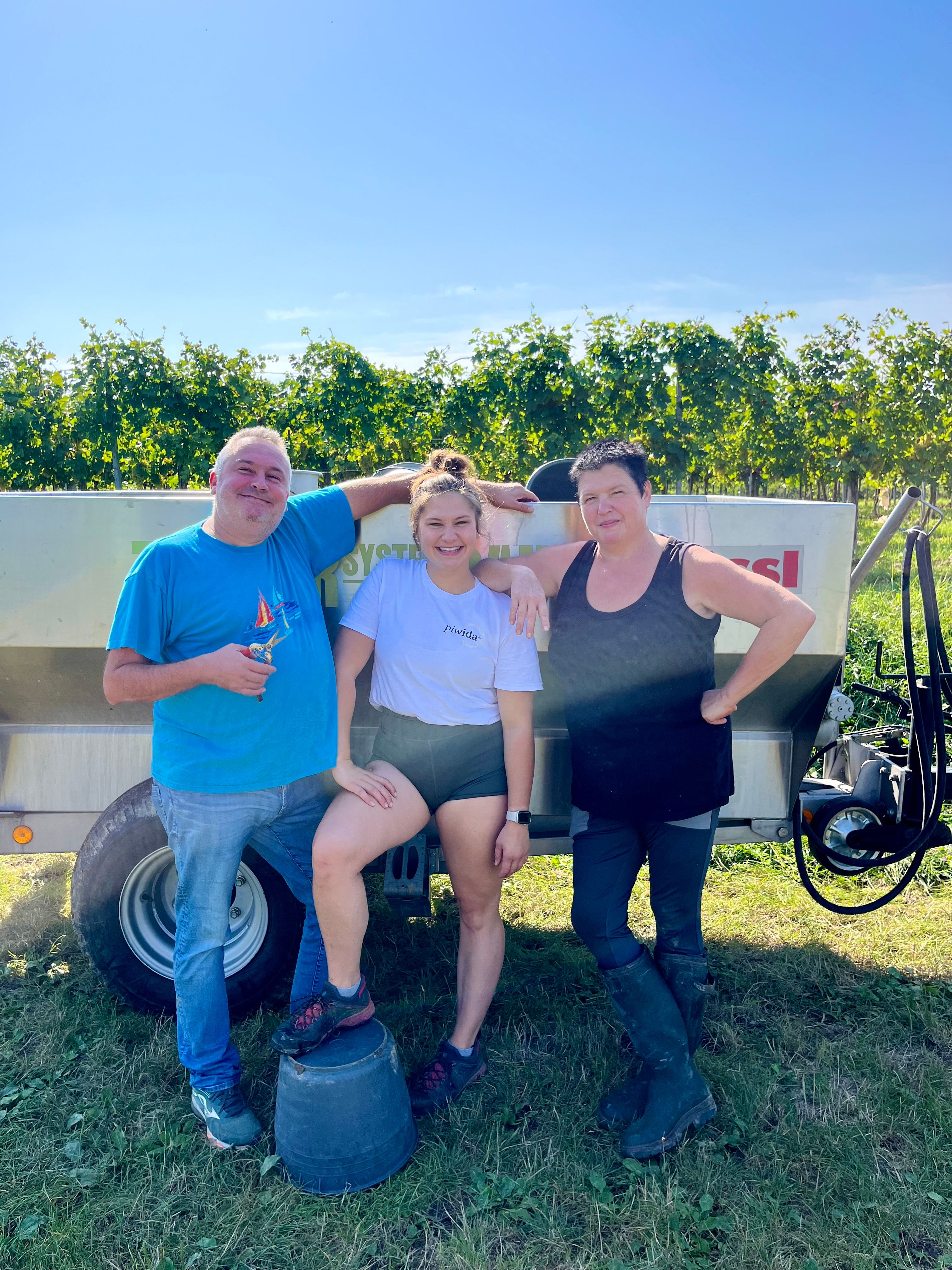 Three people are standing in front of a vineyard next to a trailer.
