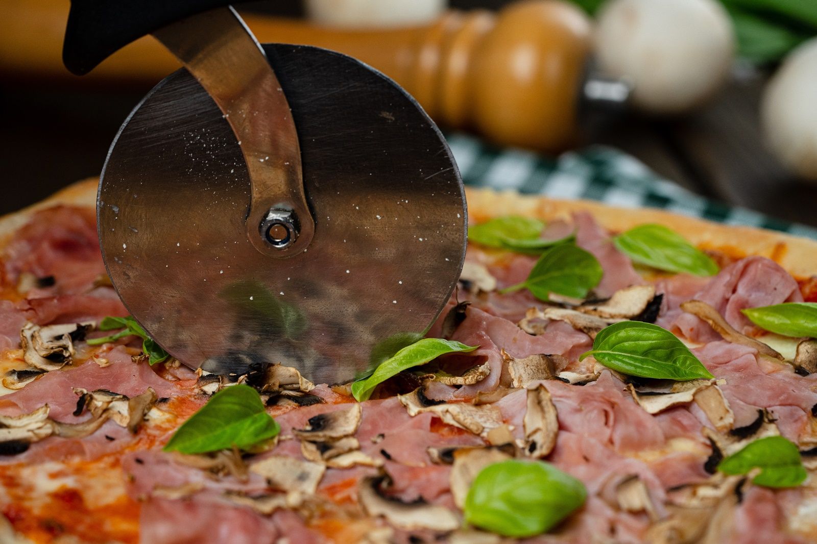 Close-up of a pizza with ham, mushrooms and basil being cut with a pizza cutter.