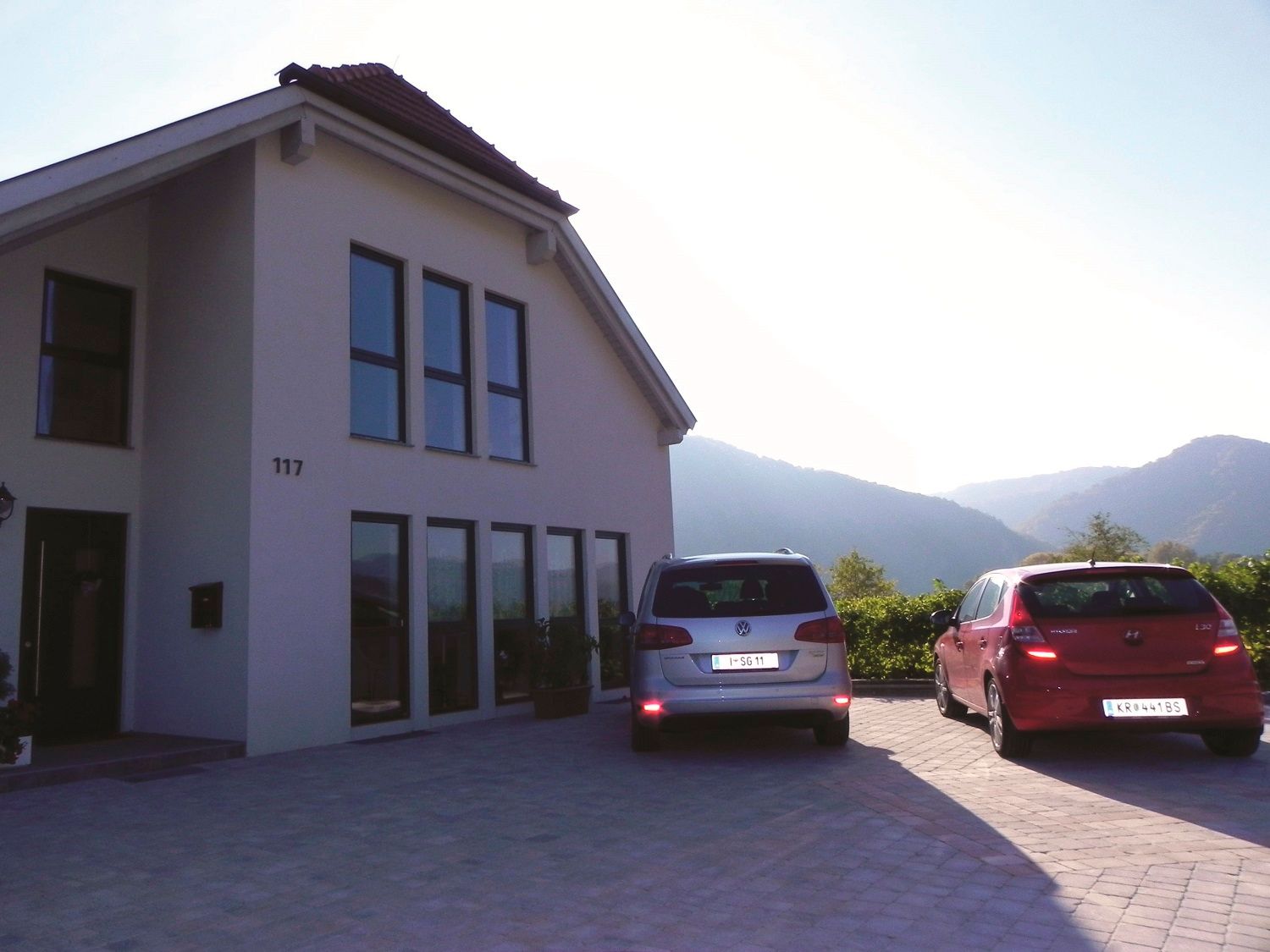 Modern house facade with two parked cars on a driveway, surrounded by hills.