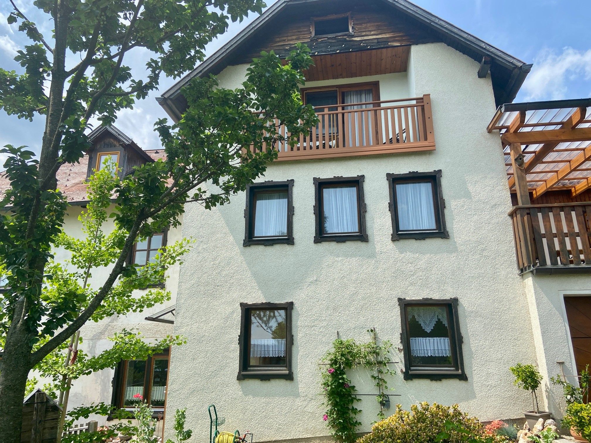 A traditional house with white walls, wooden cladding and a balcony, surrounded by green trees and plants.