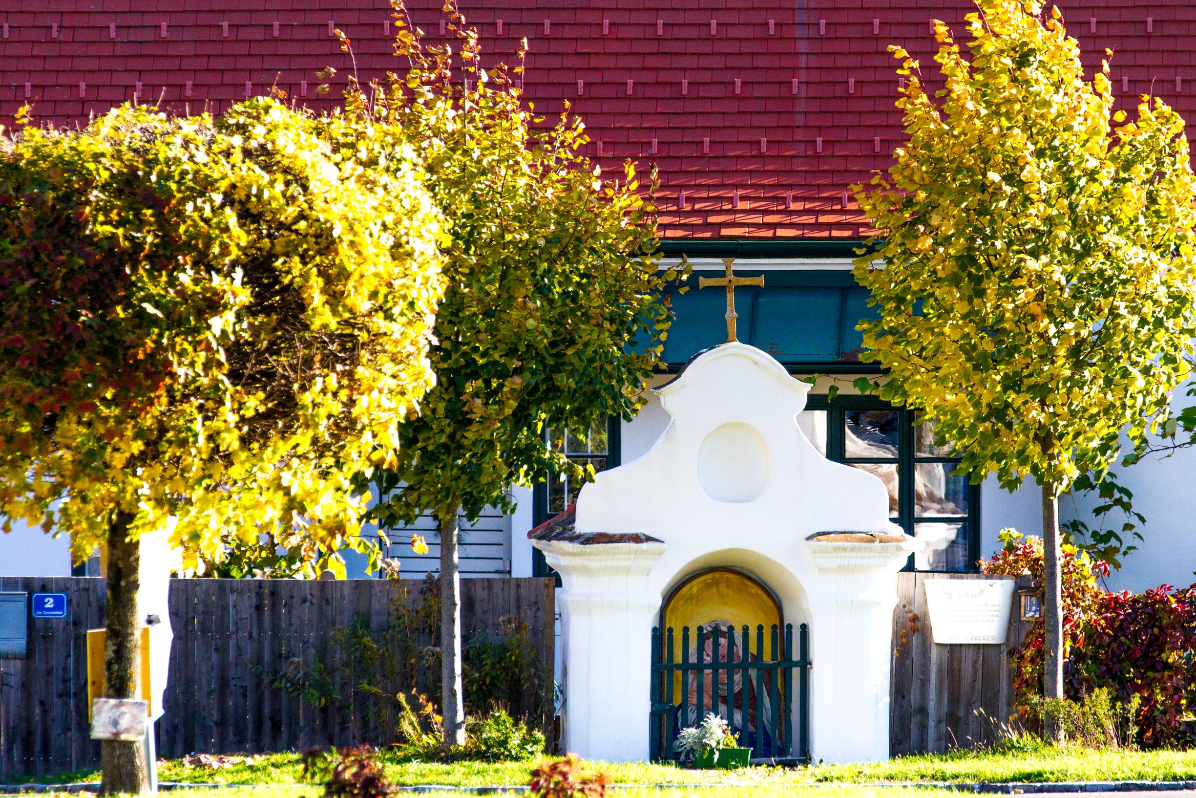 A small, white chapel building with a cross on the roof, surrounded by trees with autumn leaves.