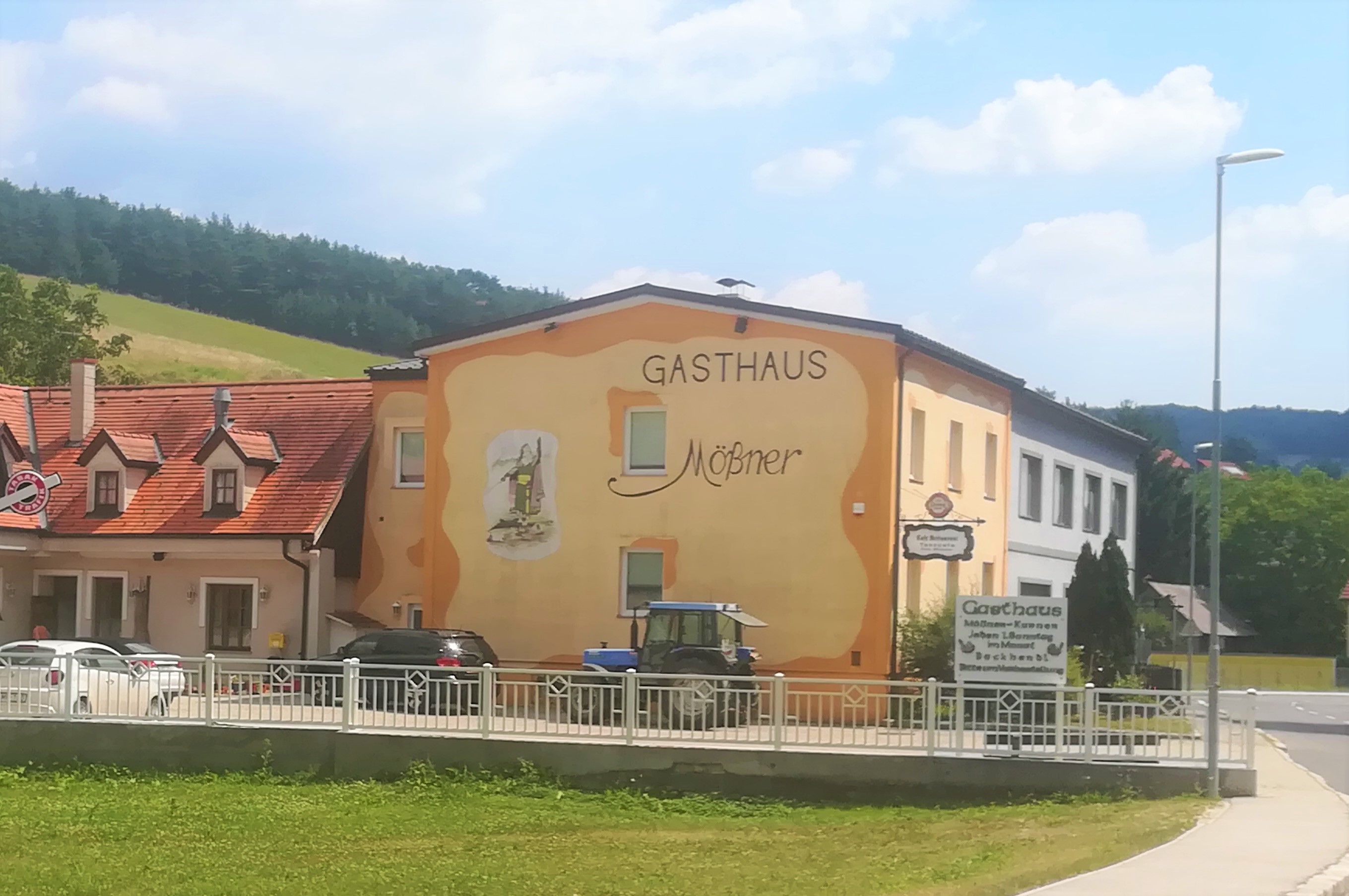 Exterior view of the Mößner inn, painted yellow-orange, in a rural setting with a blue tractor and cars in front of the building.