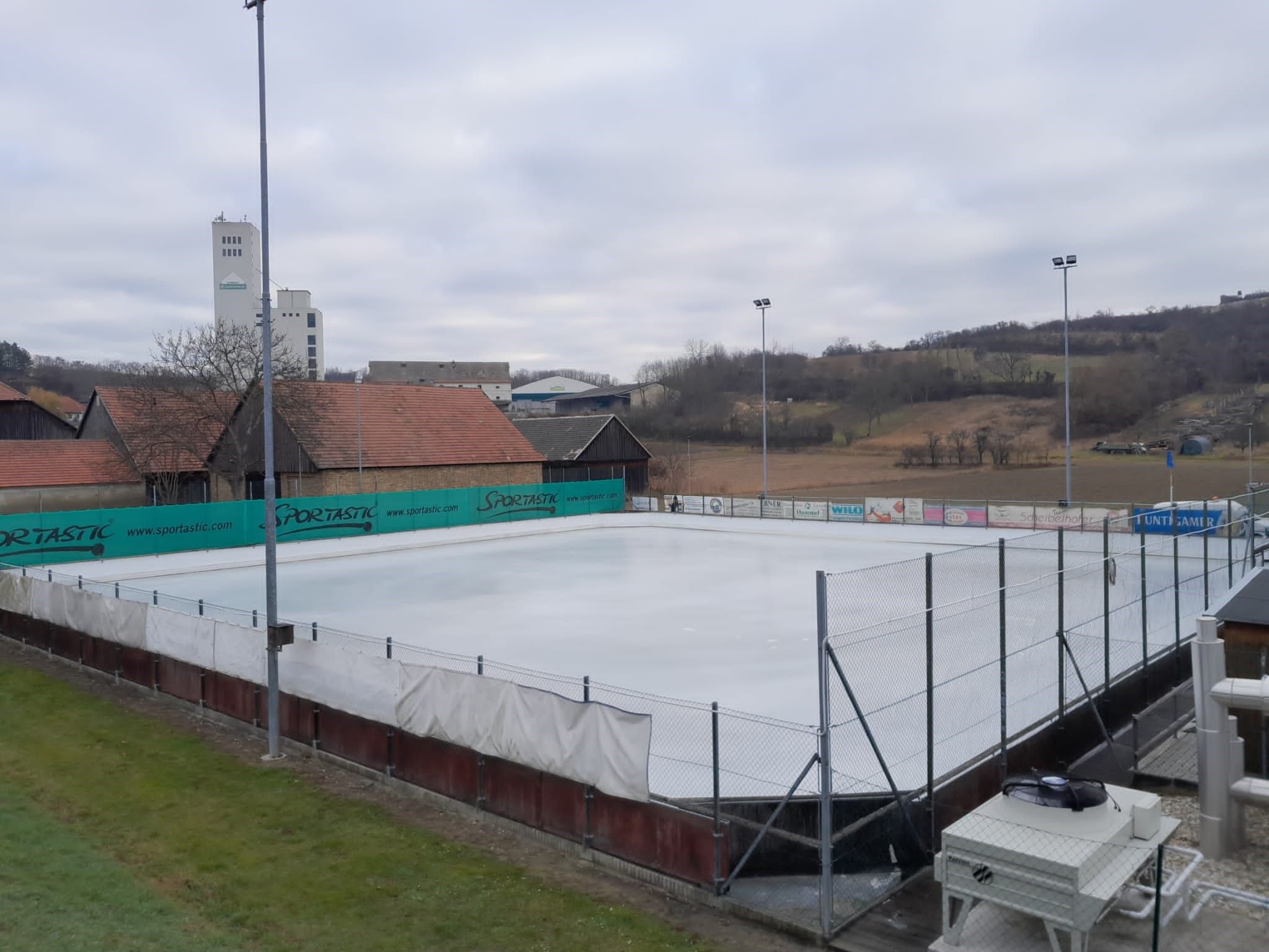 An empty ice rink with surrounding buildings and hills in the background.