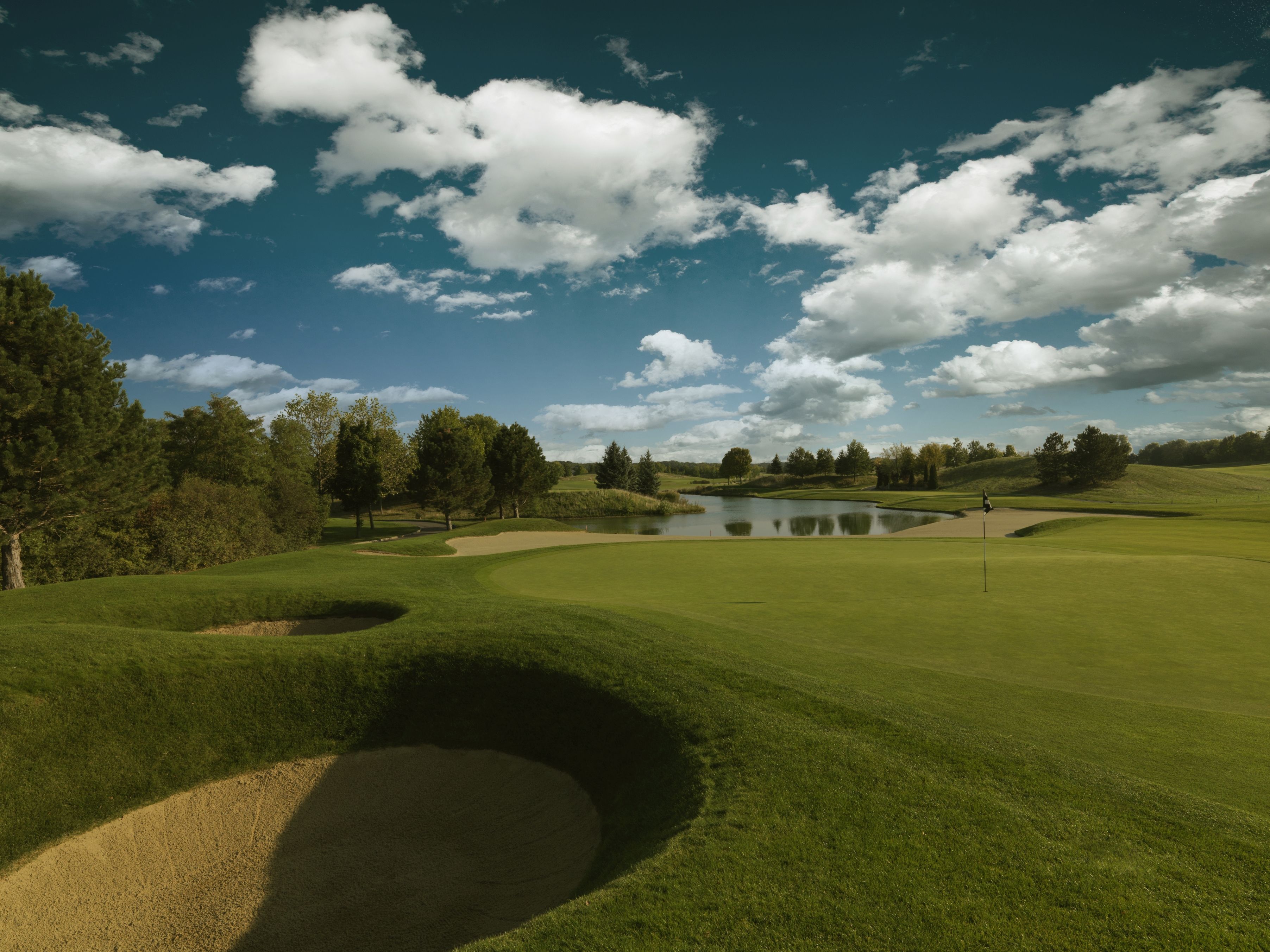 Golf course with sand bunker, green and pond under a blue sky with clouds.