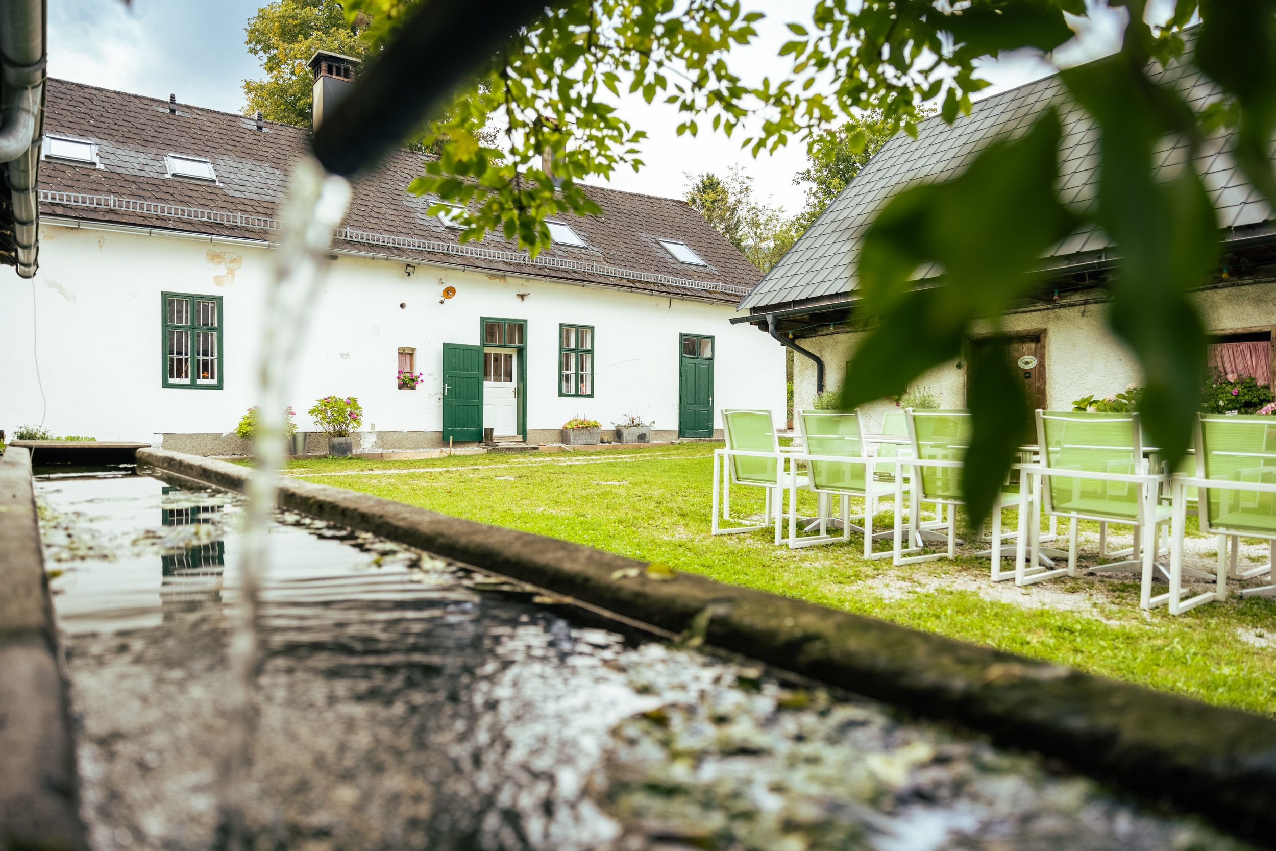 A courtyard with a white building, green lawn and garden furniture. In the foreground, water flows into a trough.