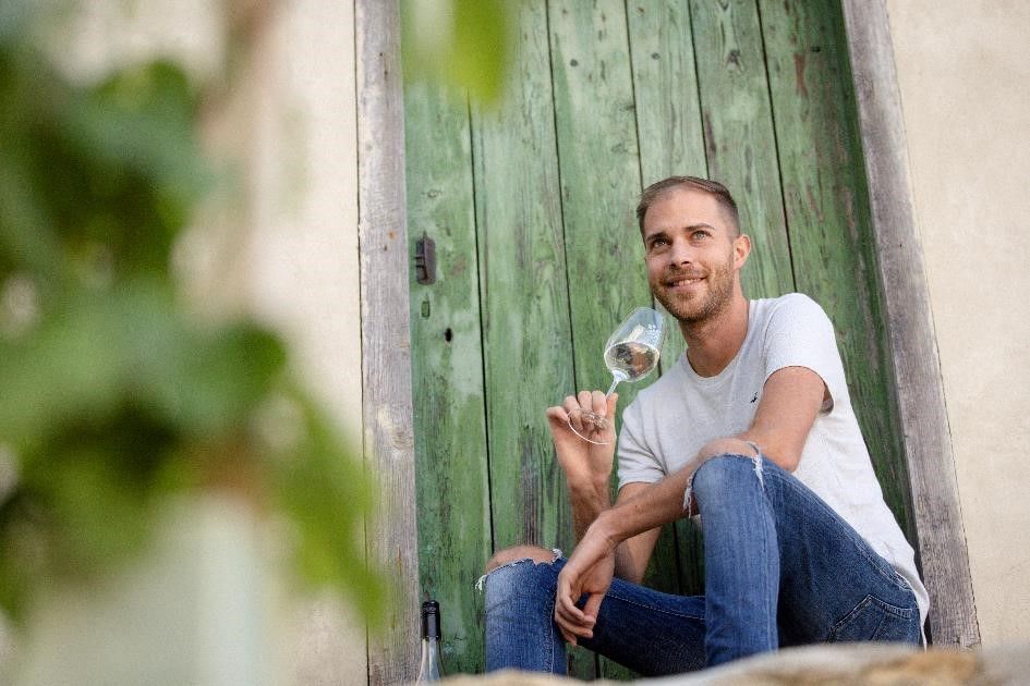A man is sitting in front of a green wooden door, holding a wine glass in his hand.