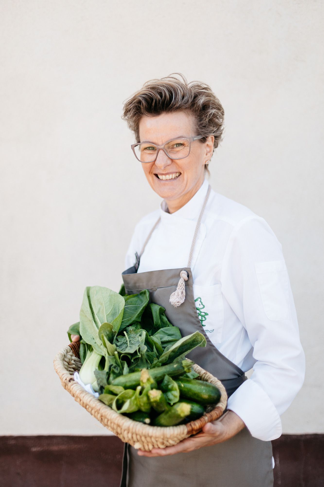 A smiling woman in chef's clothes holds a basket of fresh vegetables.