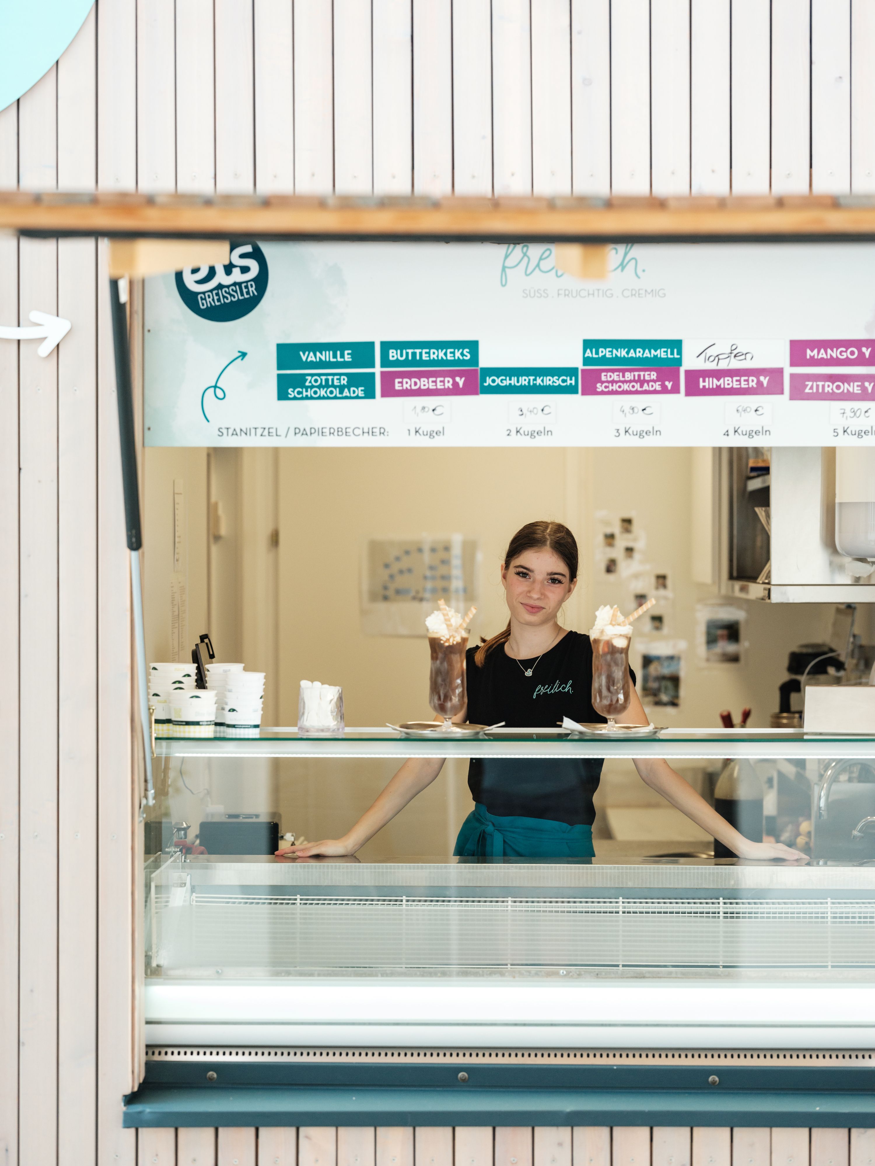 A woman stands behind an ice cream counter in an ice cream store, surrounded by different types of ice cream on a sign.
