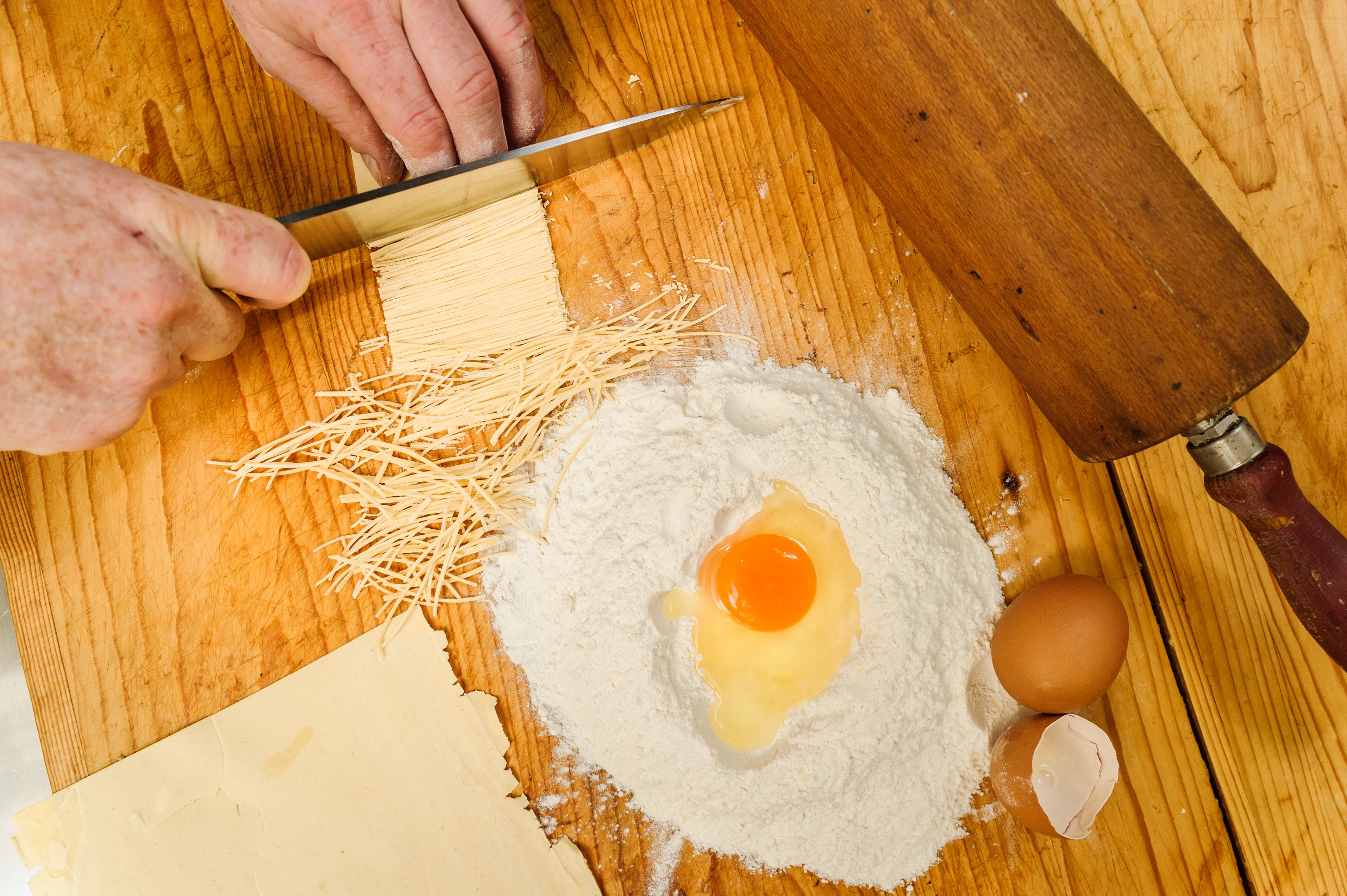 Close-up of hands cutting pasta dough, with flour, egg and rolling pin on a wooden table.