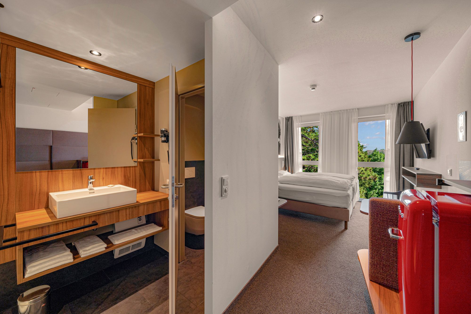 Interior view of a modern hotel room with bathroom, bed and red fridge.