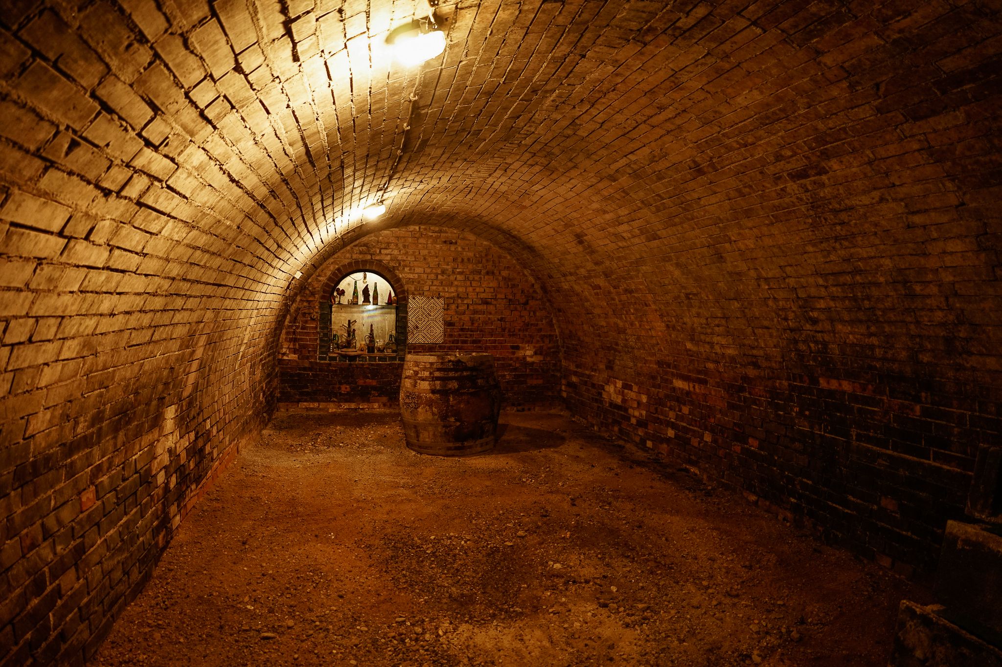 A vaulted brick cellar with a barrel and an illuminated shelf in the background.