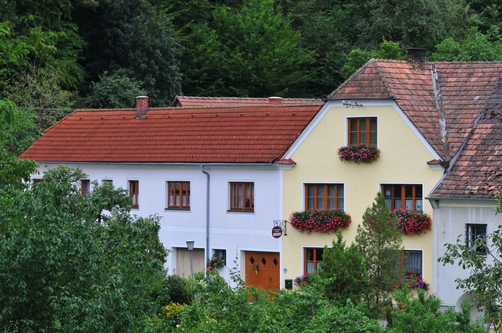 A traditional house with red roof tiles and flower boxes in front of the windows, surrounded by green vegetation.