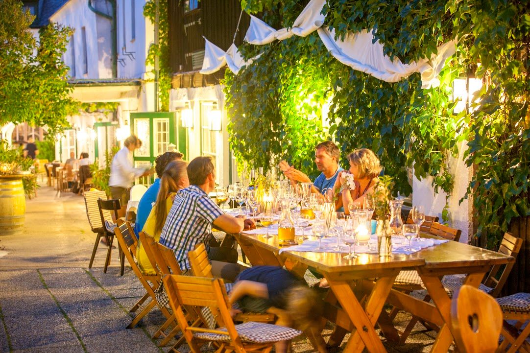 People sit at a long table outside, surrounded by plants and lights.