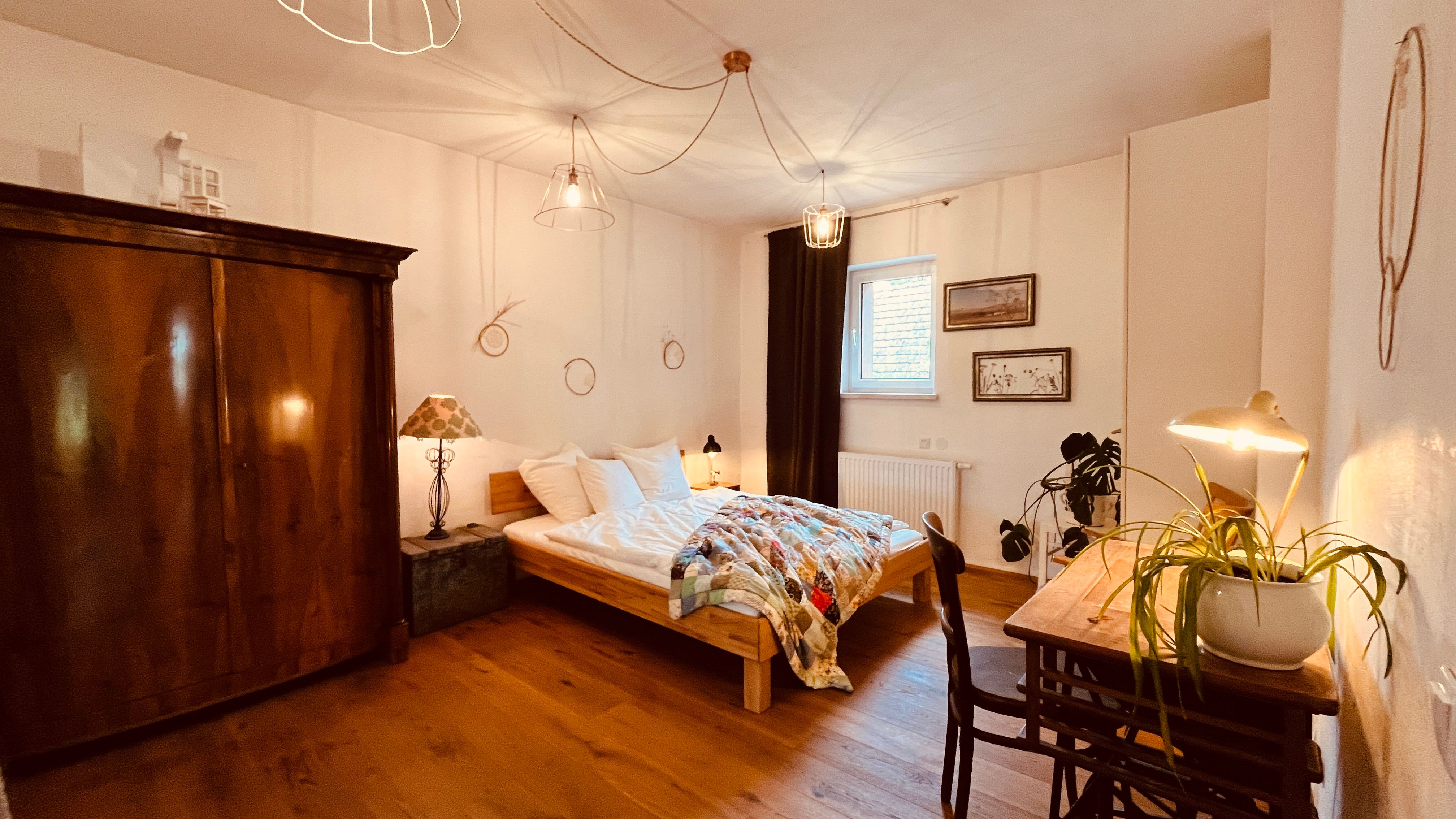 Cozy bedroom with wooden floor, double bed, antique wardrobe, desk with lamp and plant, as well as decorative elements on the walls.
