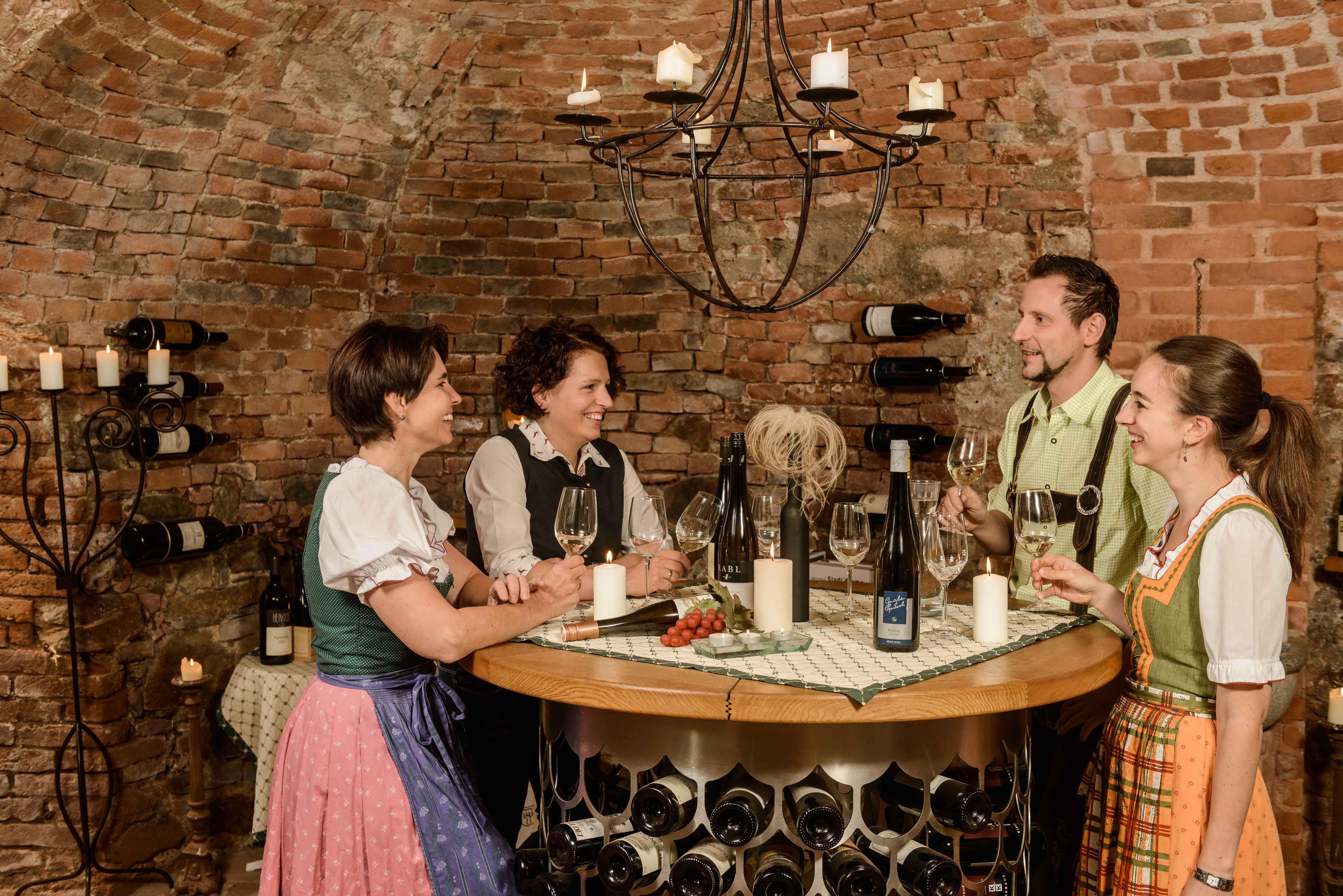 Four people in traditional dress are standing in a wine cellar holding wine glasses. They are laughing and chatting at a round table.