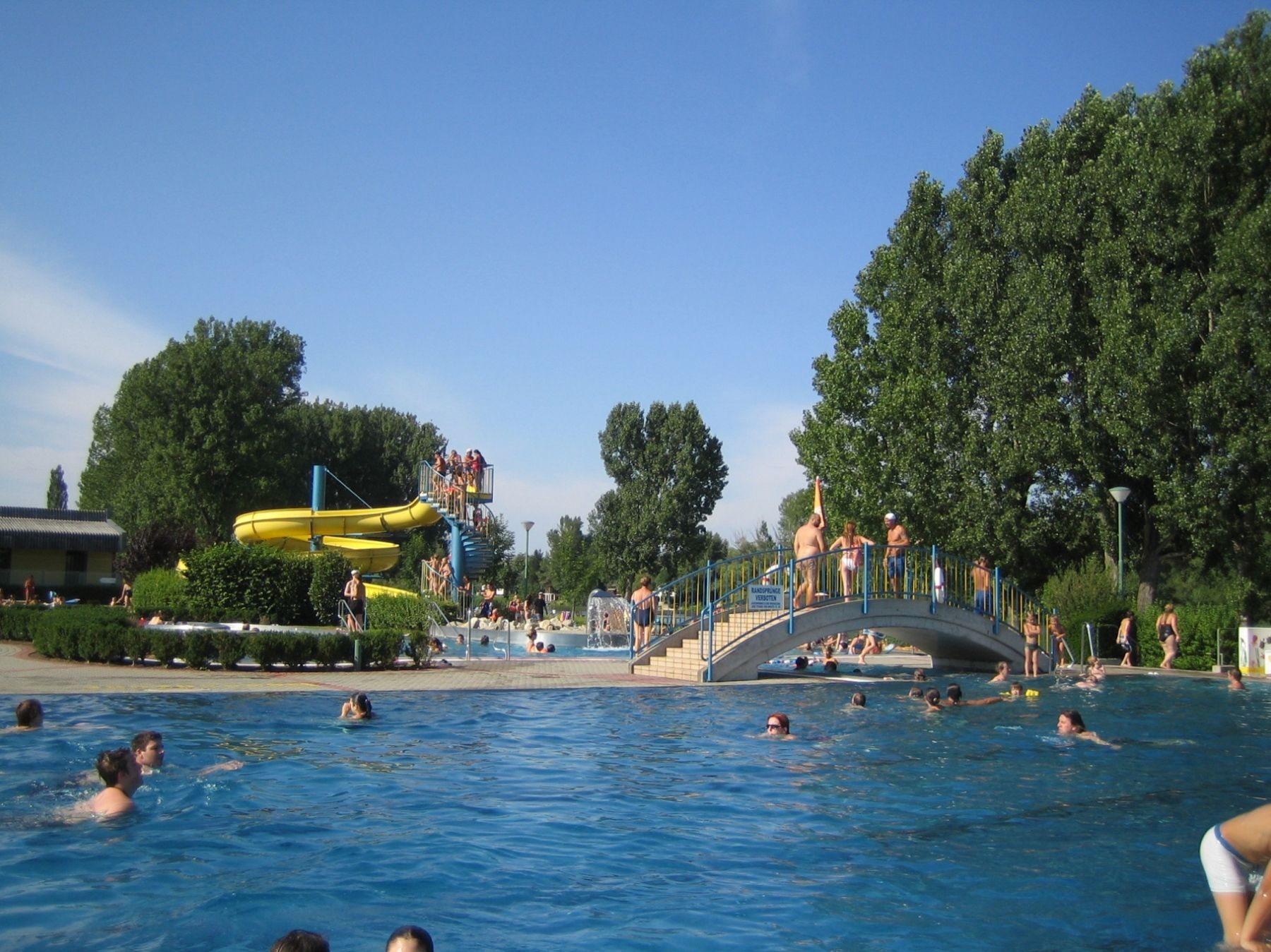 Outdoor pool with water slide and bridge, lots of people in the water.
