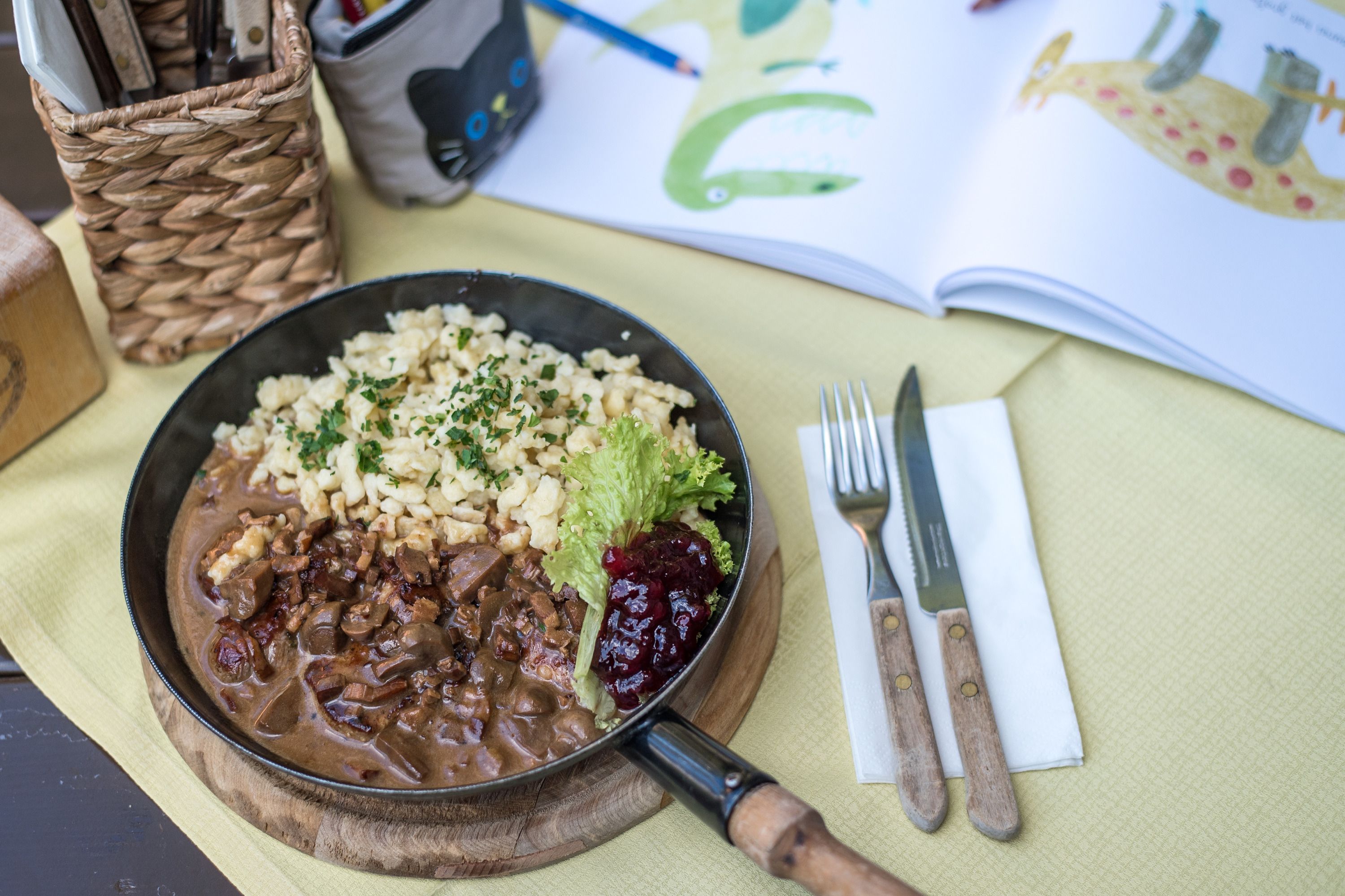 A plate of spaetzle, meat and cranberries on a table with cutlery and an open book.