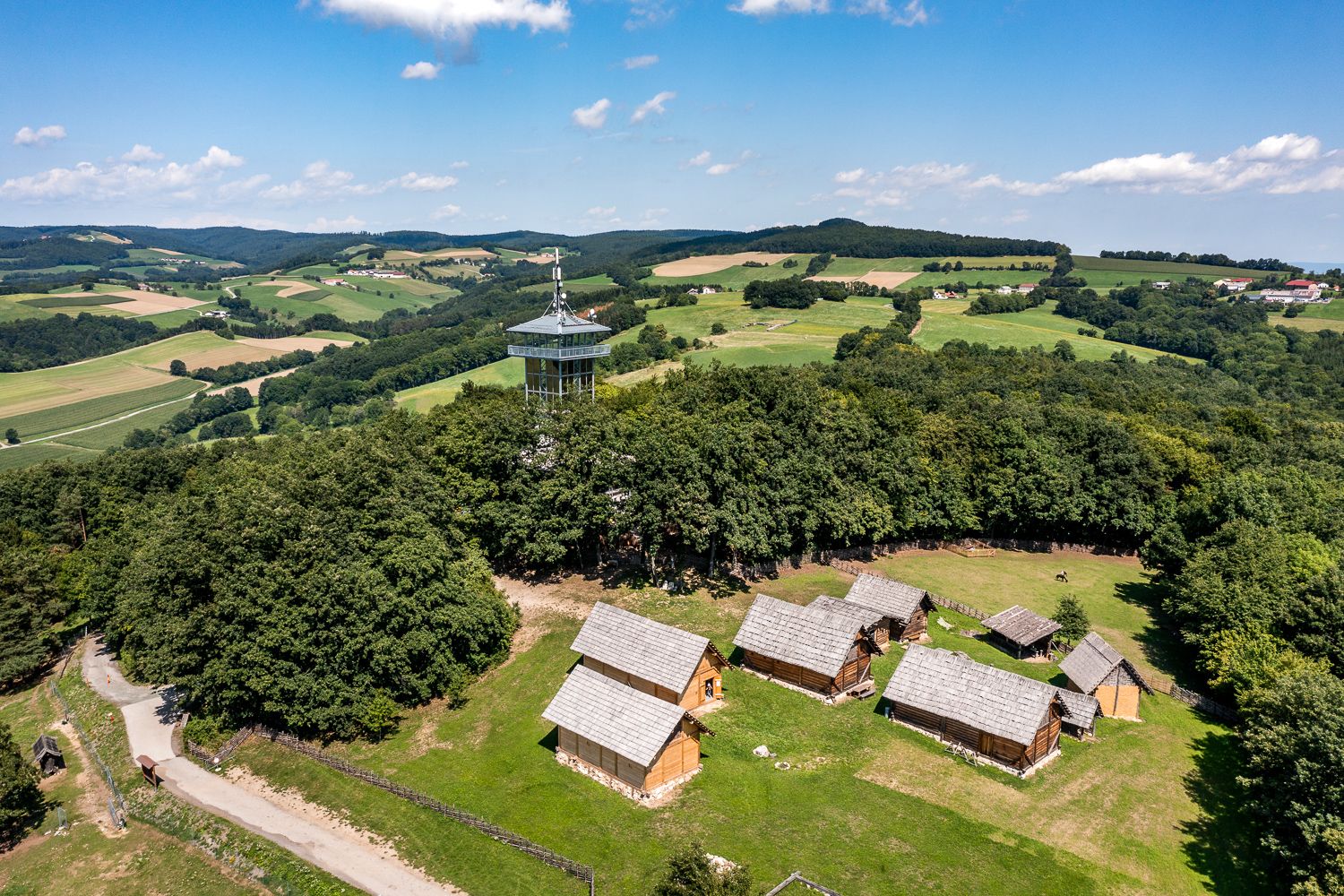 Aerial view of the Celtic village of Schwarzenbach with wooden houses and a lookout tower, surrounded by green fields and forests.