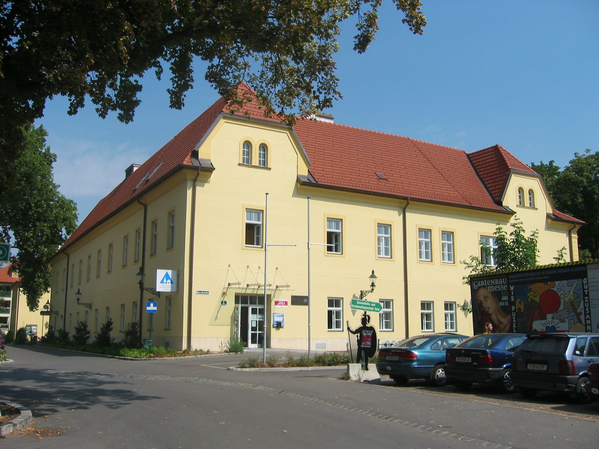 Exterior view of a yellow building with a red roof, Junges Hotel Tulln, surrounded by trees and parked cars.