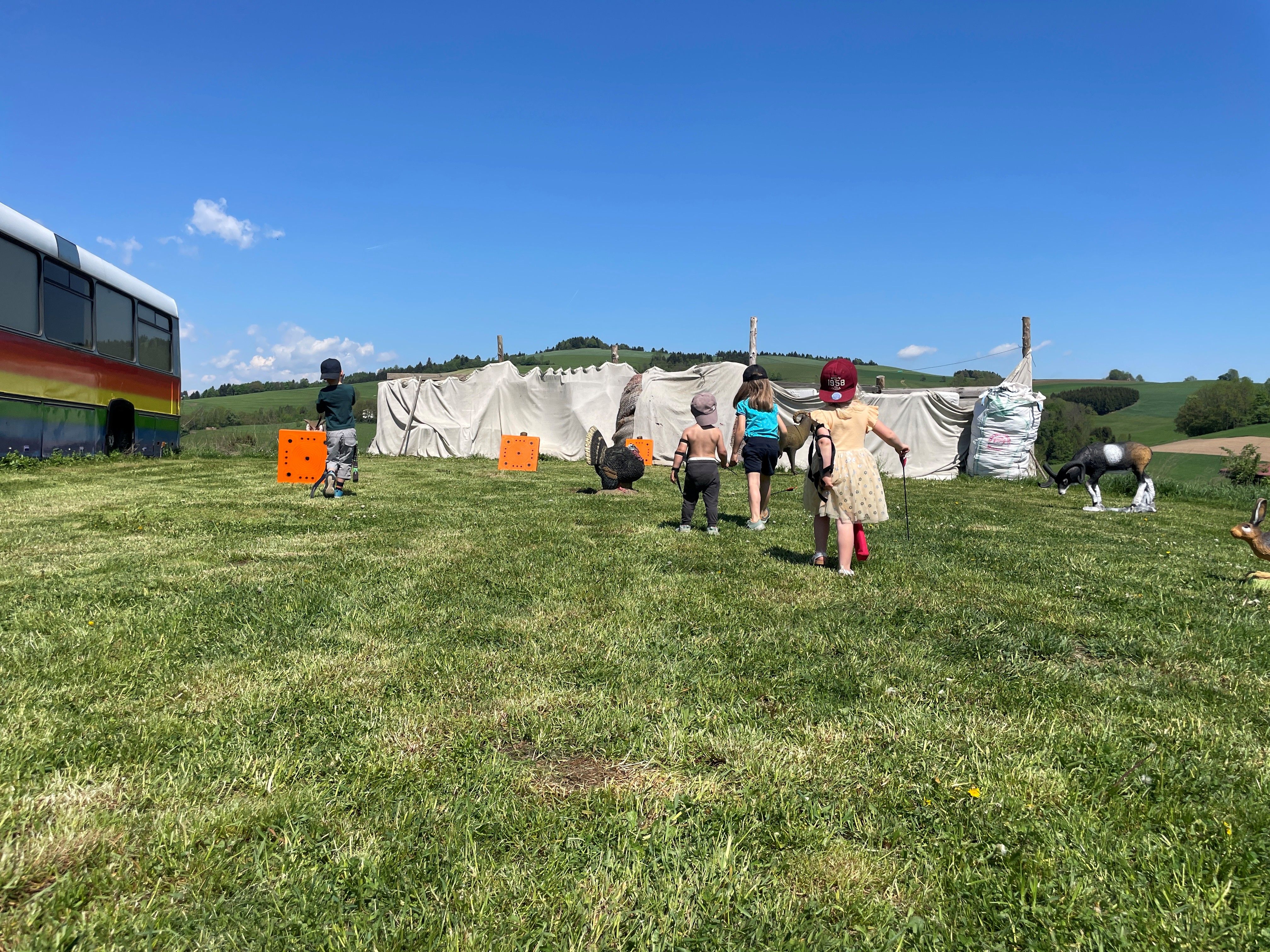 Children play on a meadow with archery targets and a colorful bus in the background.
