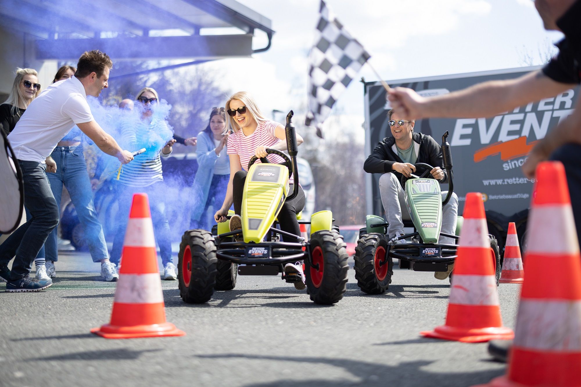 Participants ride pedal tractors at an event with blue smoke and spectators.