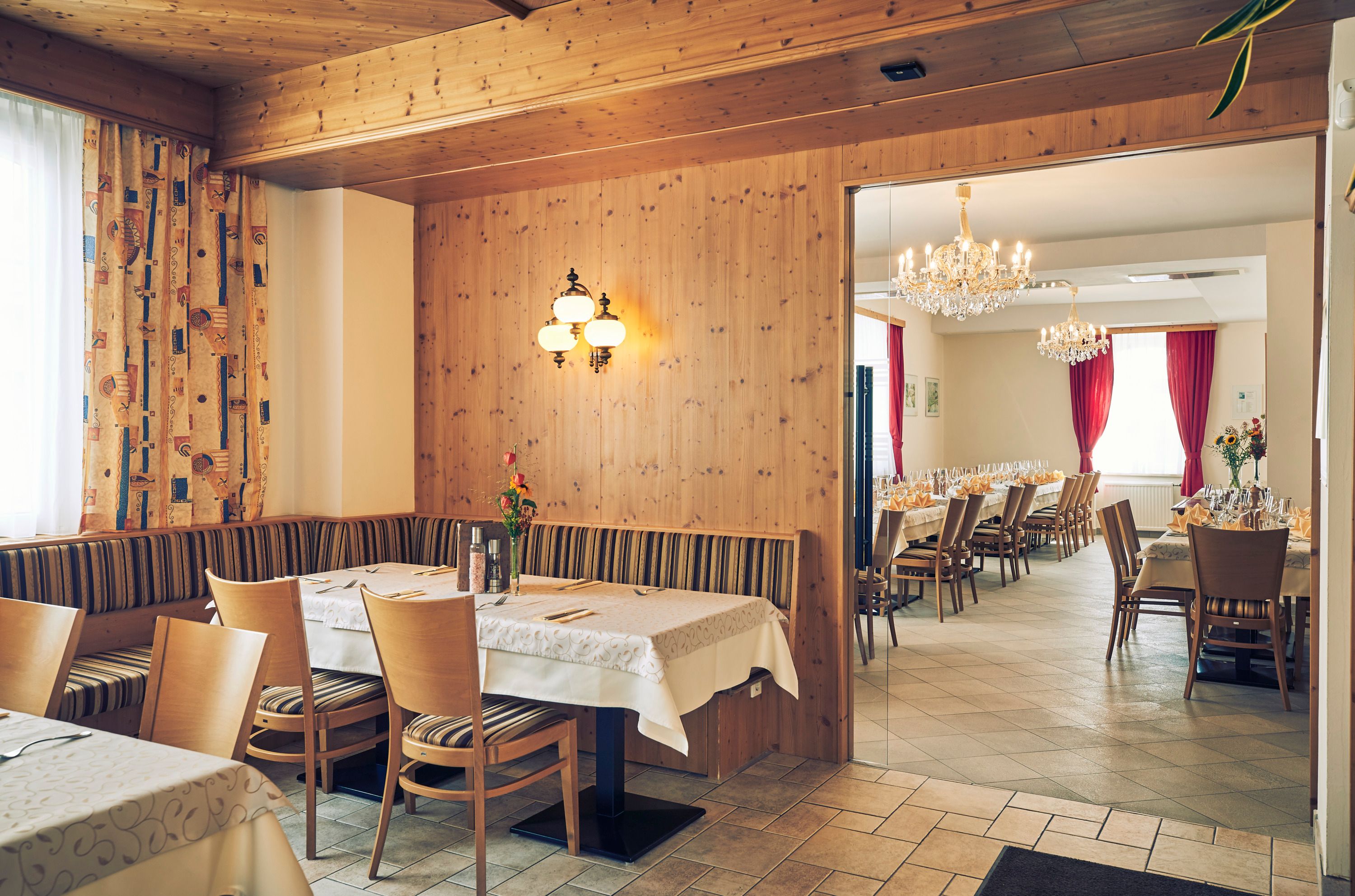 A cozy guest room with wooden walls and an adjoining ballroom with chandeliers and laid tables.