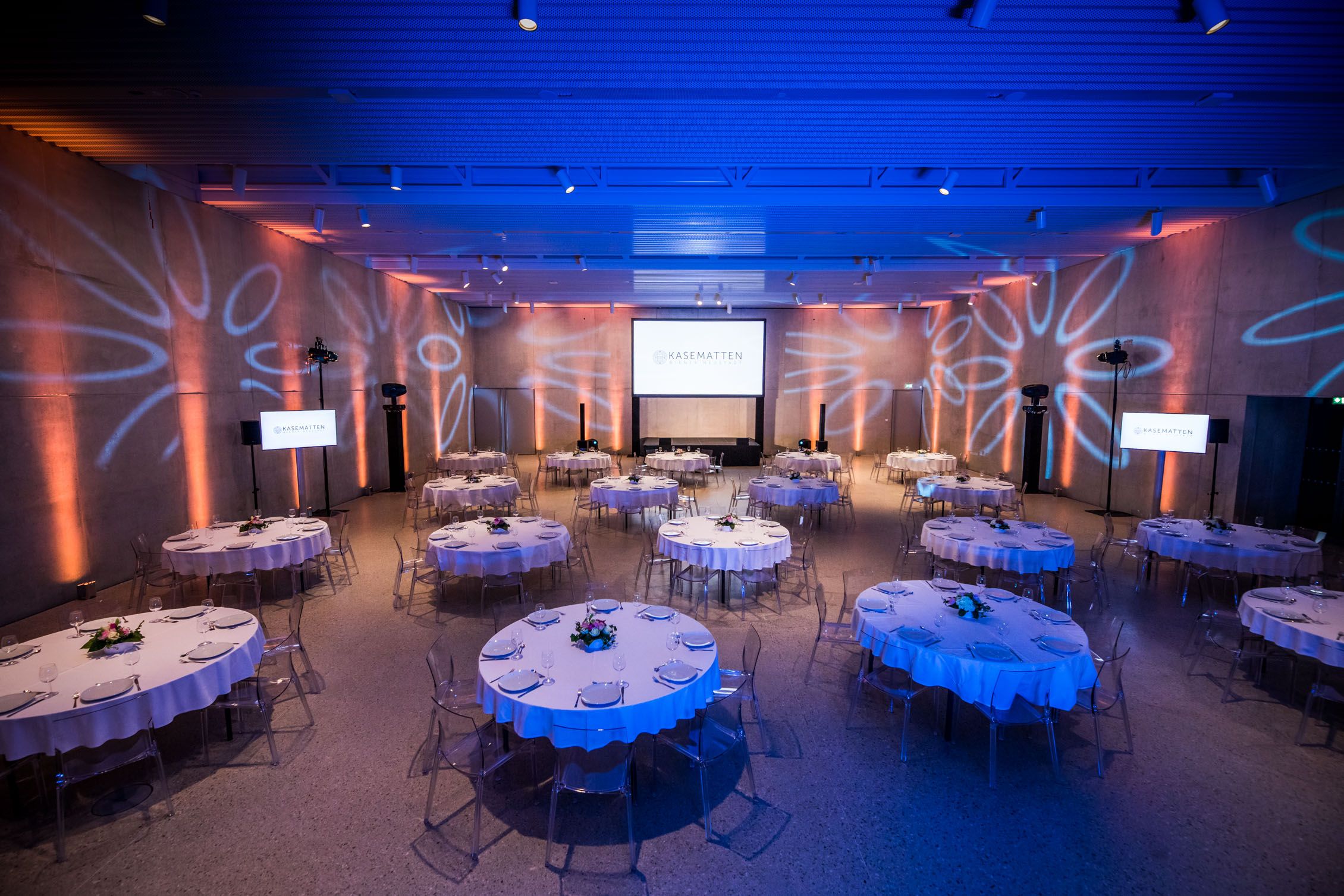 An elegant function room with round, laid tables and blue lighting.