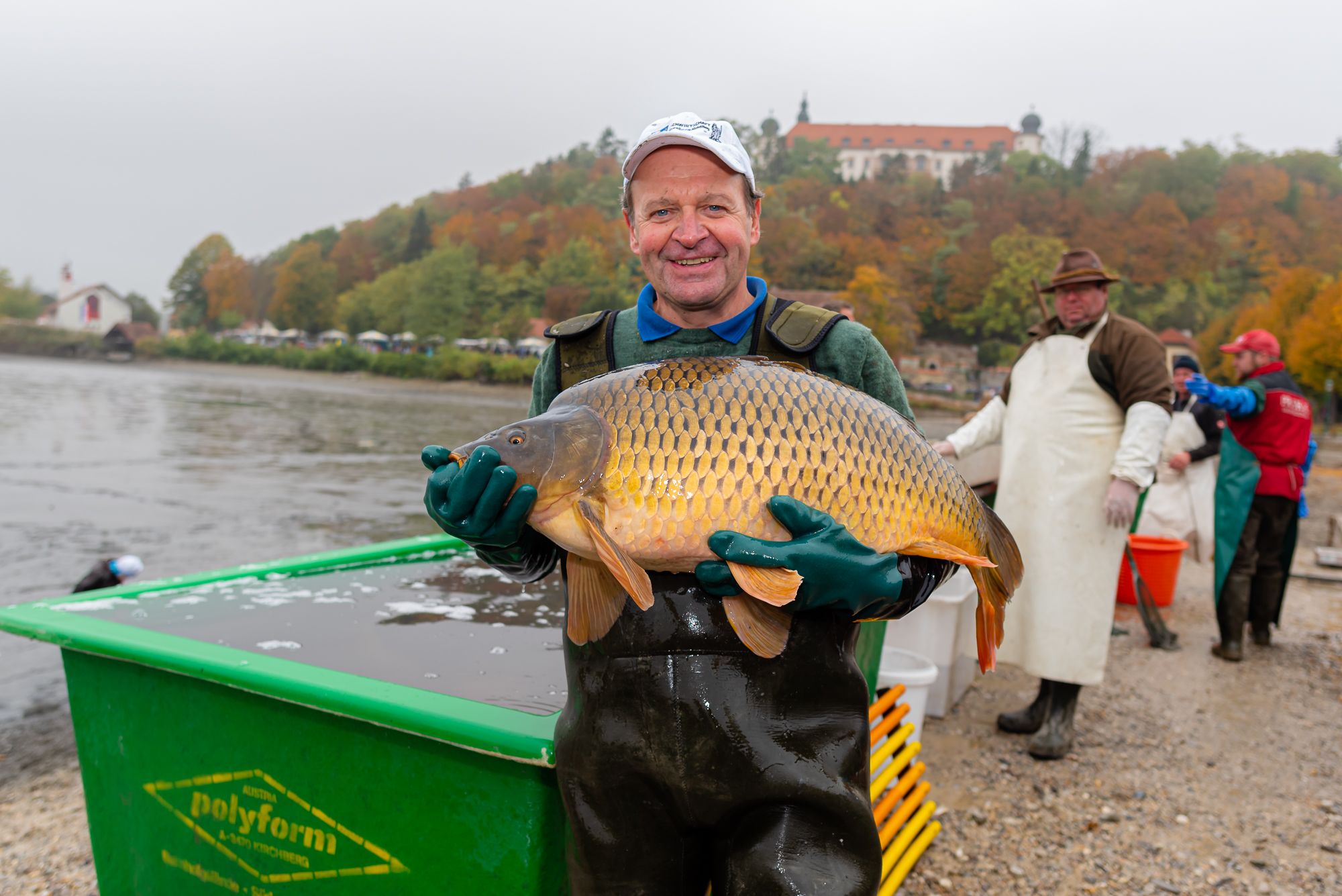 A man holds a large carp in front of a pond. Other people and a building on a hill can be seen in the background.