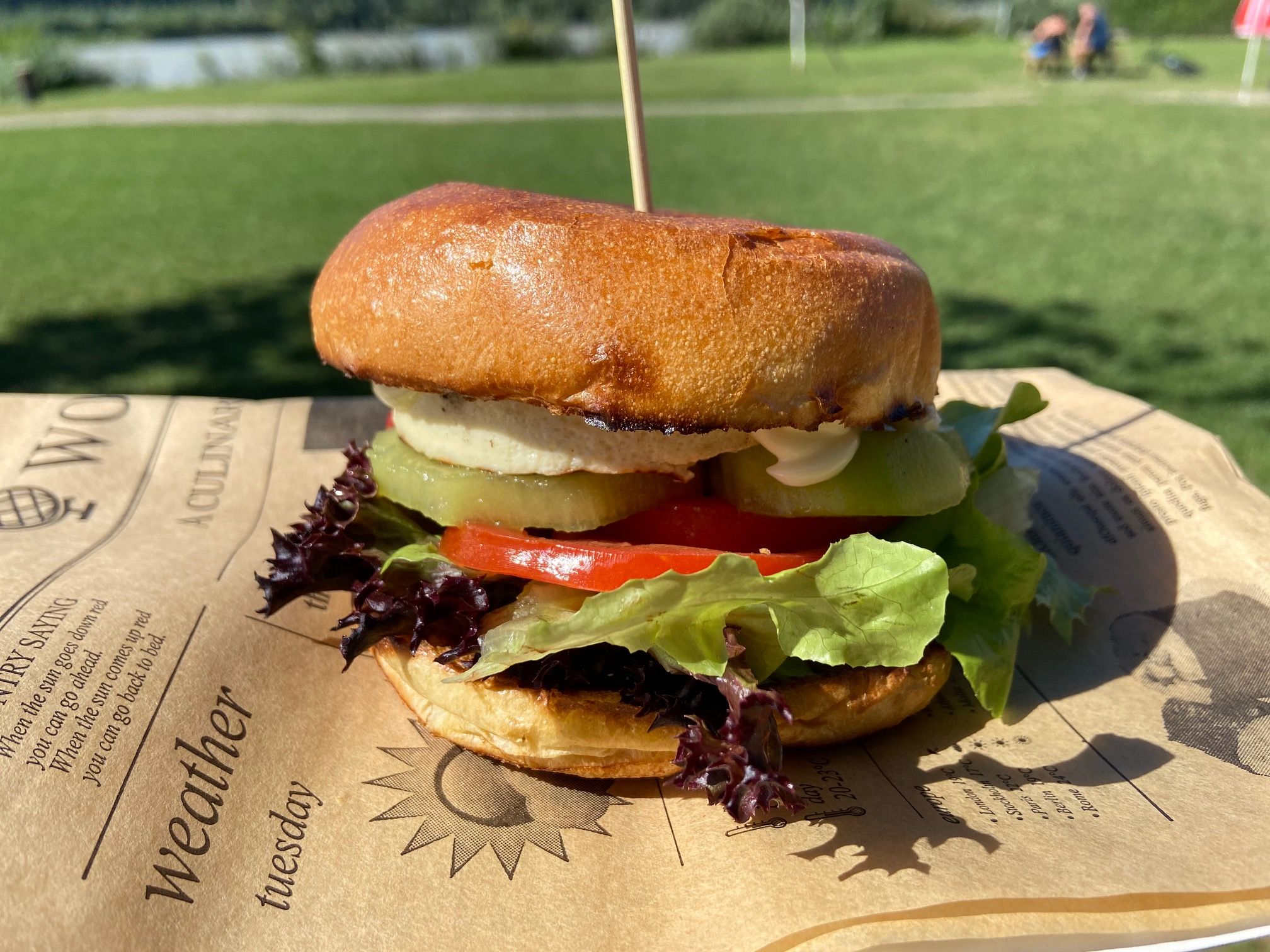A burger with lettuce, tomatoes and gherkins on a tray outside.