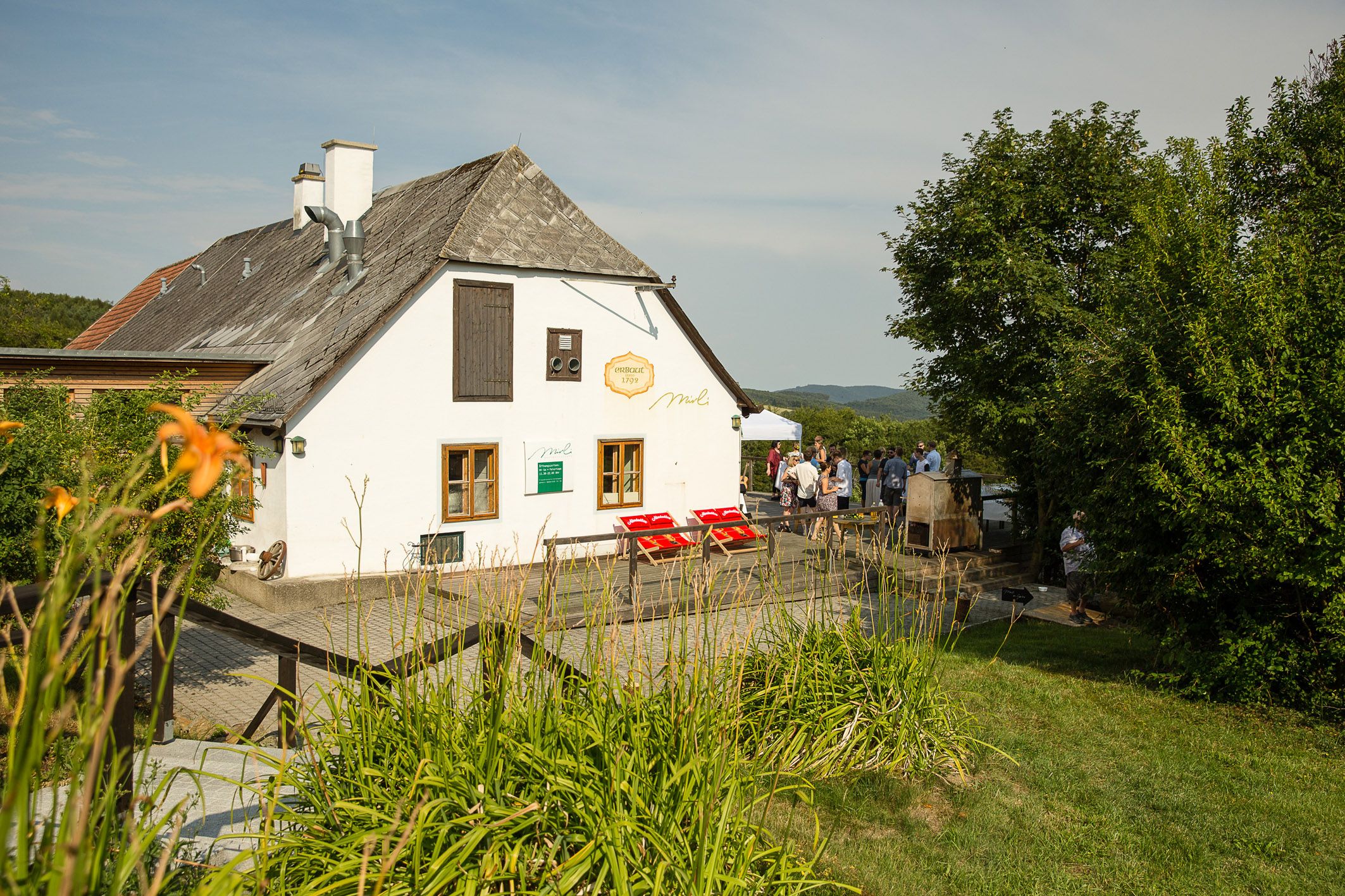 White country house with shingle roof, surrounded by green landscape and people in the background.