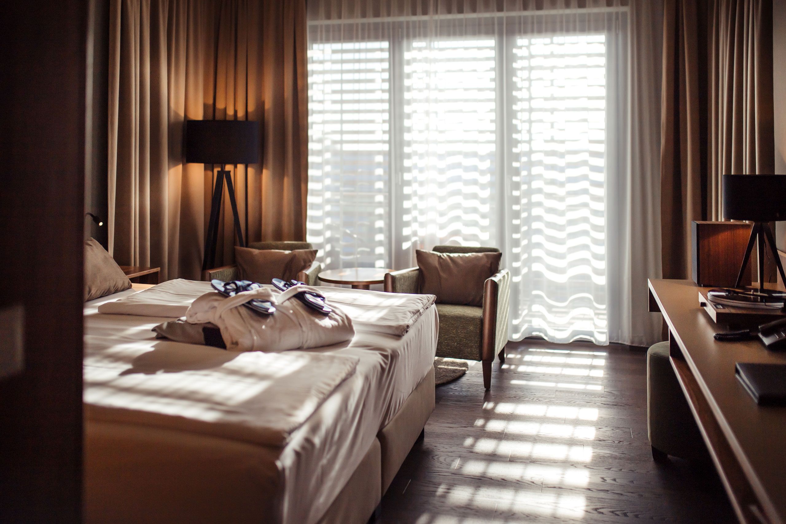 Hotel room with double bed, bathrobes, two armchairs and a table in front of a large window with curtains.