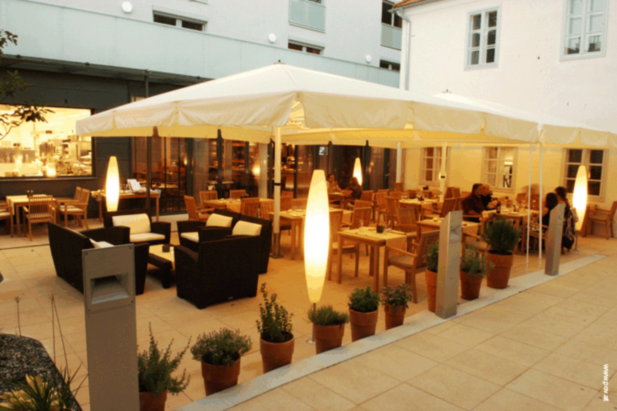 An illuminated guest garden with tables, chairs and plants in pots, surrounded by buildings.