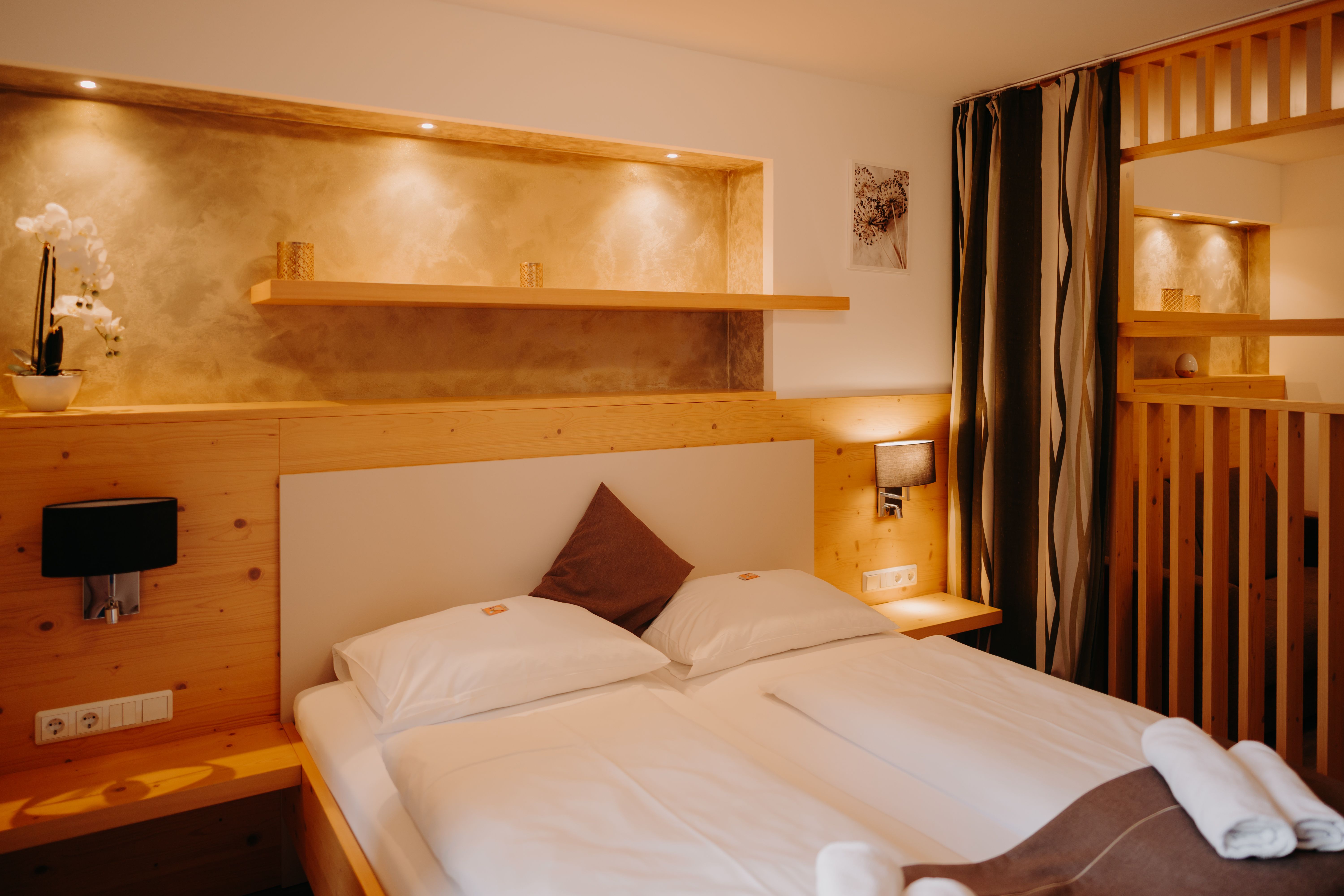 A cozy hotel room with a double bed, light-colored wood paneling, a wooden room divider, and a curtain. There are electrical outlets and light switches next to the bed. Above the bed is a wall niche that is illuminated and painted in an earthy gold tone.