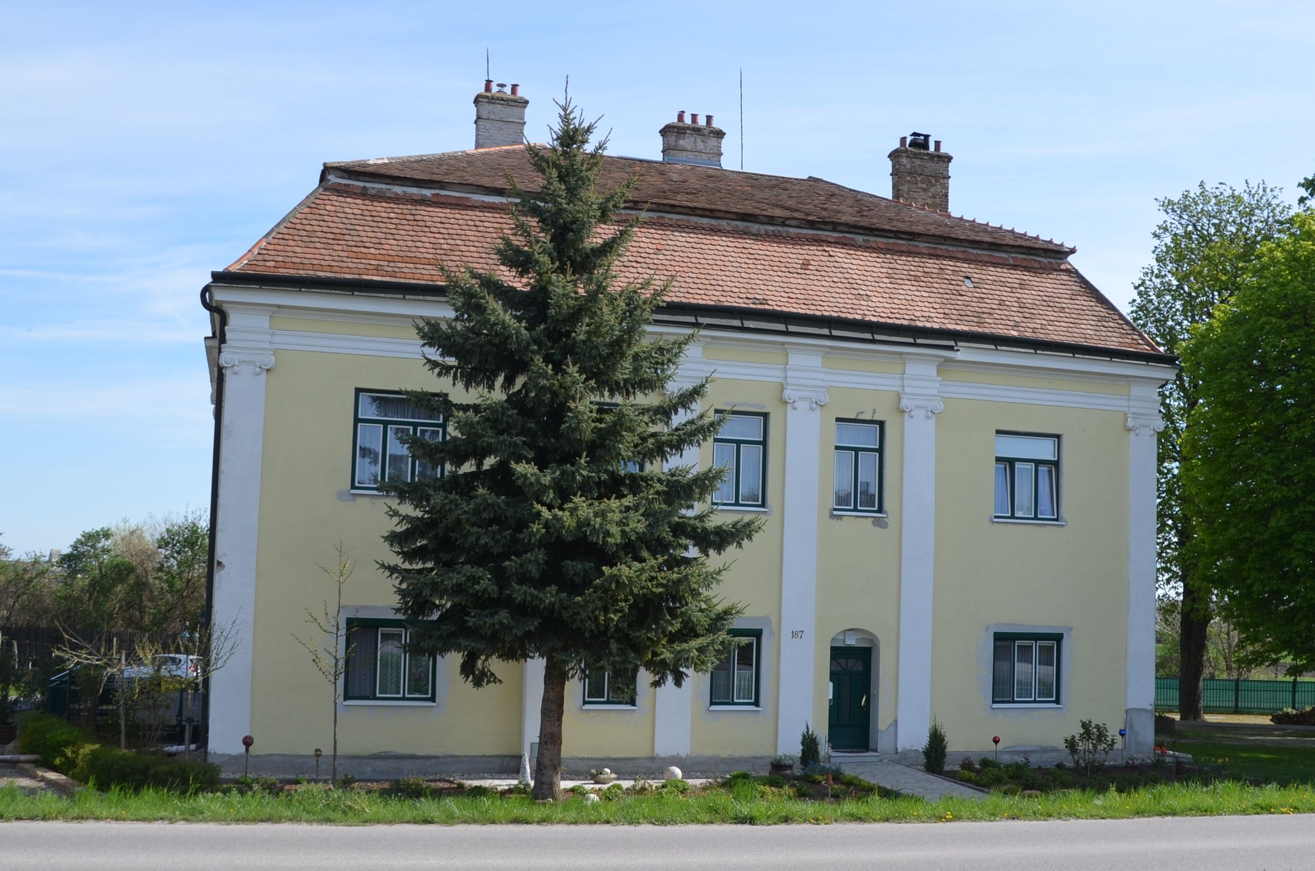 Yellow two-storey building with red tiled roof and green door, surrounded by trees and a small garden.