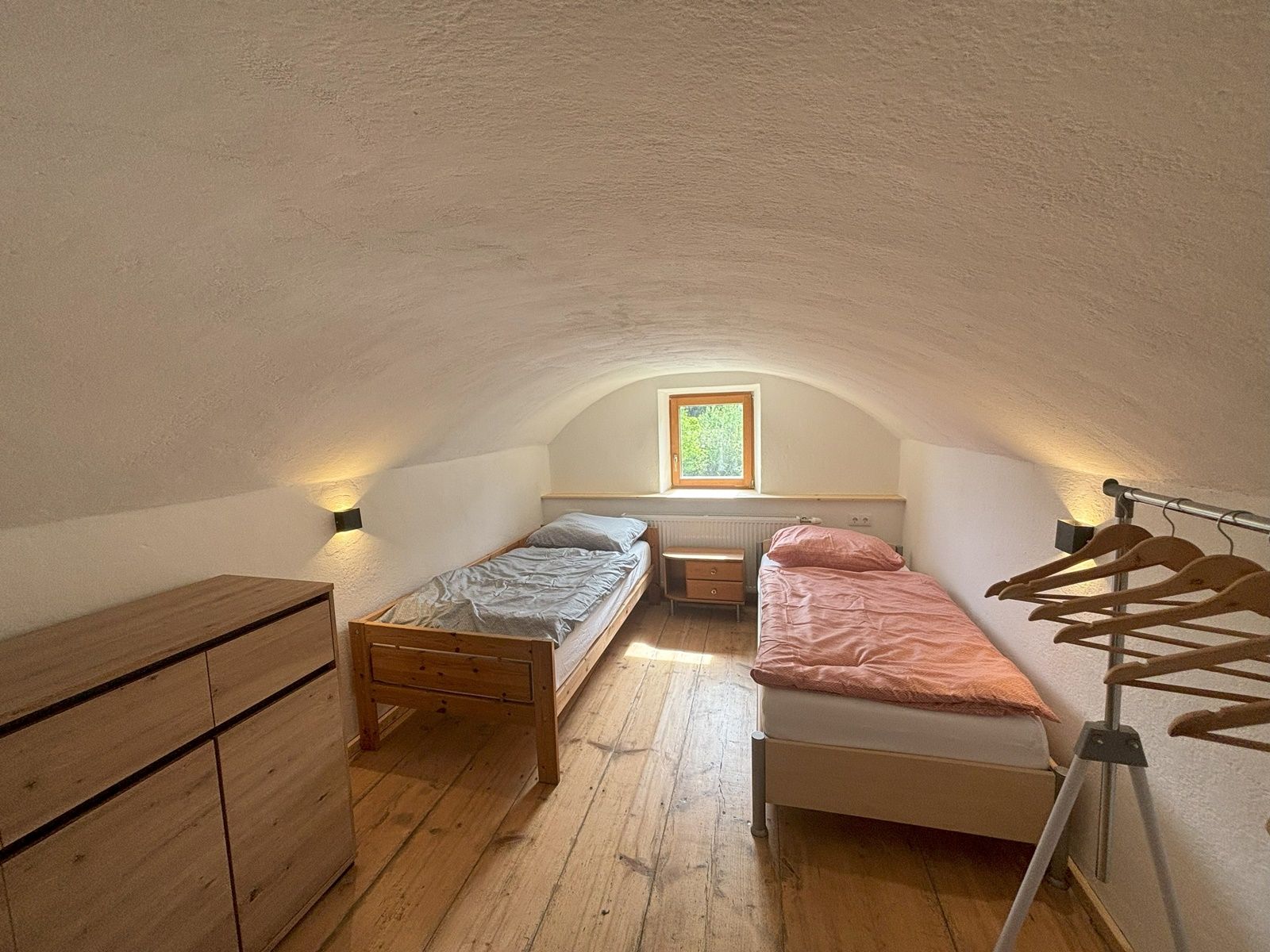 A simple double room with two single beds, a chest of drawers and a clothes rack. A small window lets in natural light.
