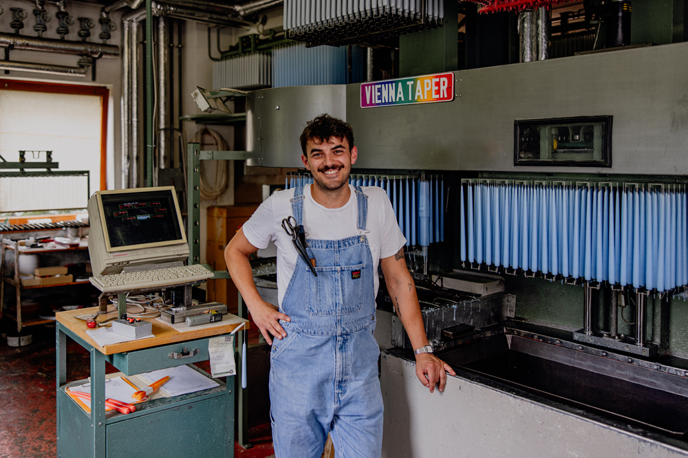 A man in dungarees stands smiling in front of a candle-dunk installation with blue candles.