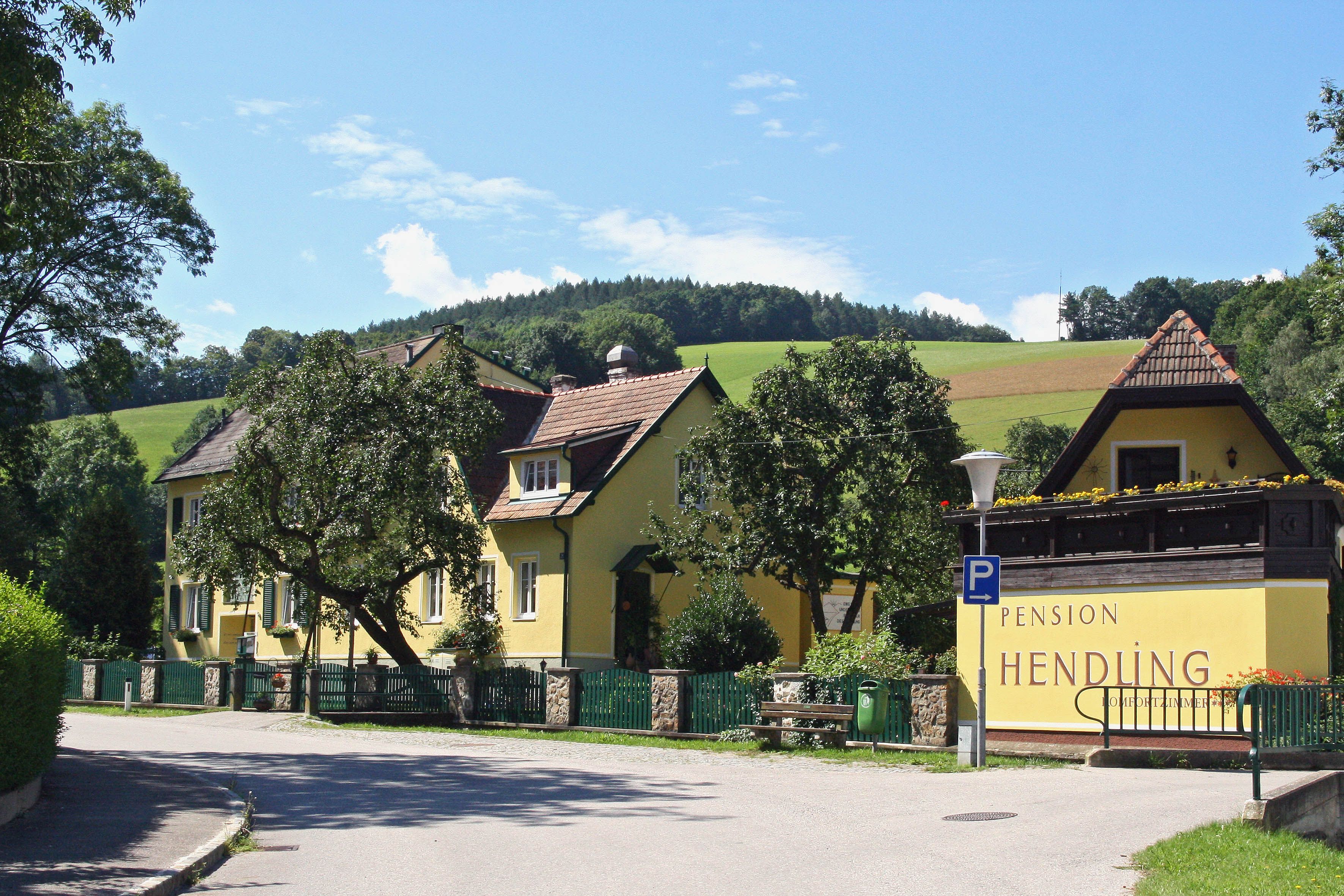 Yellow building of the Pension Hendling in a rural setting with green hills in the background.