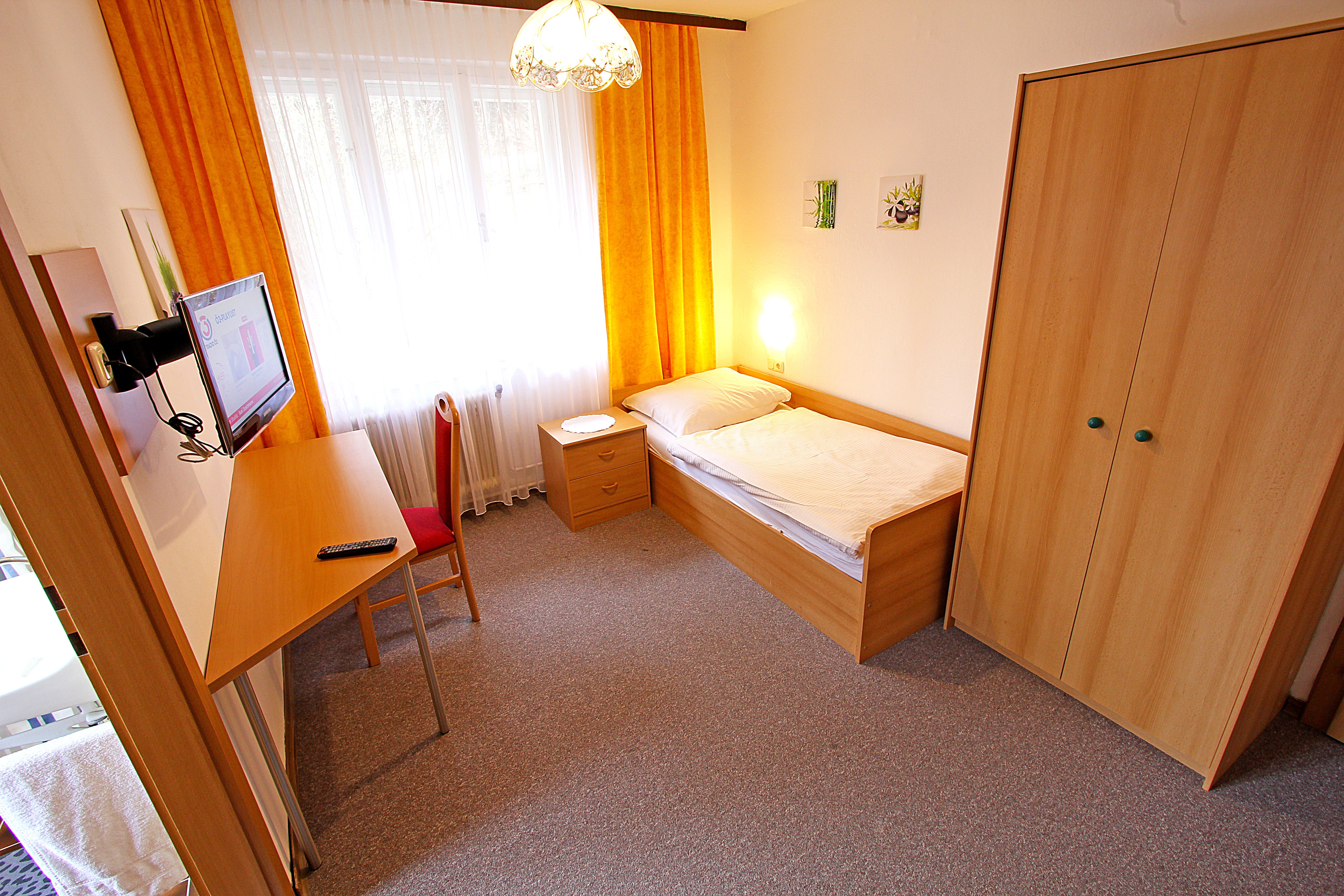 A simply furnished single room with bed, desk, chair, closet and TV. Yellow curtains and carpeted floor.