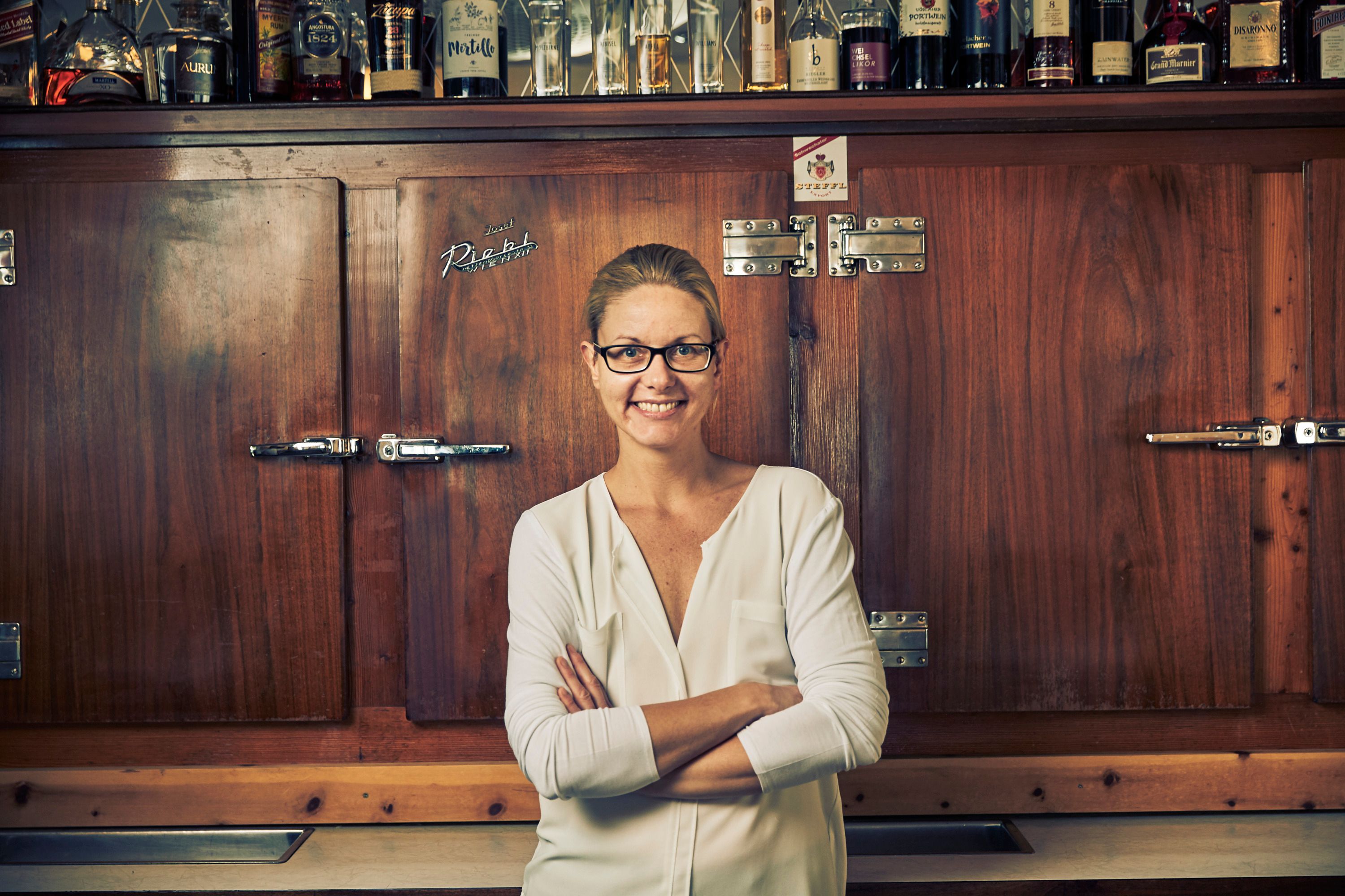 A woman with glasses stands smiling in front of a wooden cabinet with bottles on it.