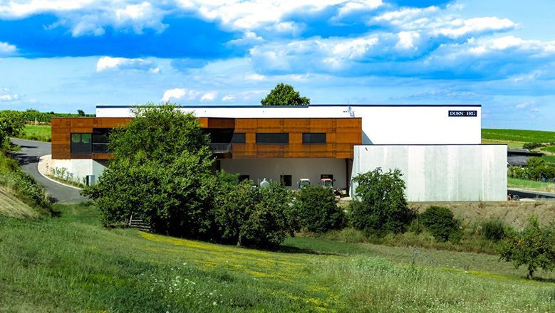Modern winery buildings with brown and white facades, surrounded by a green landscape and blue sky.
