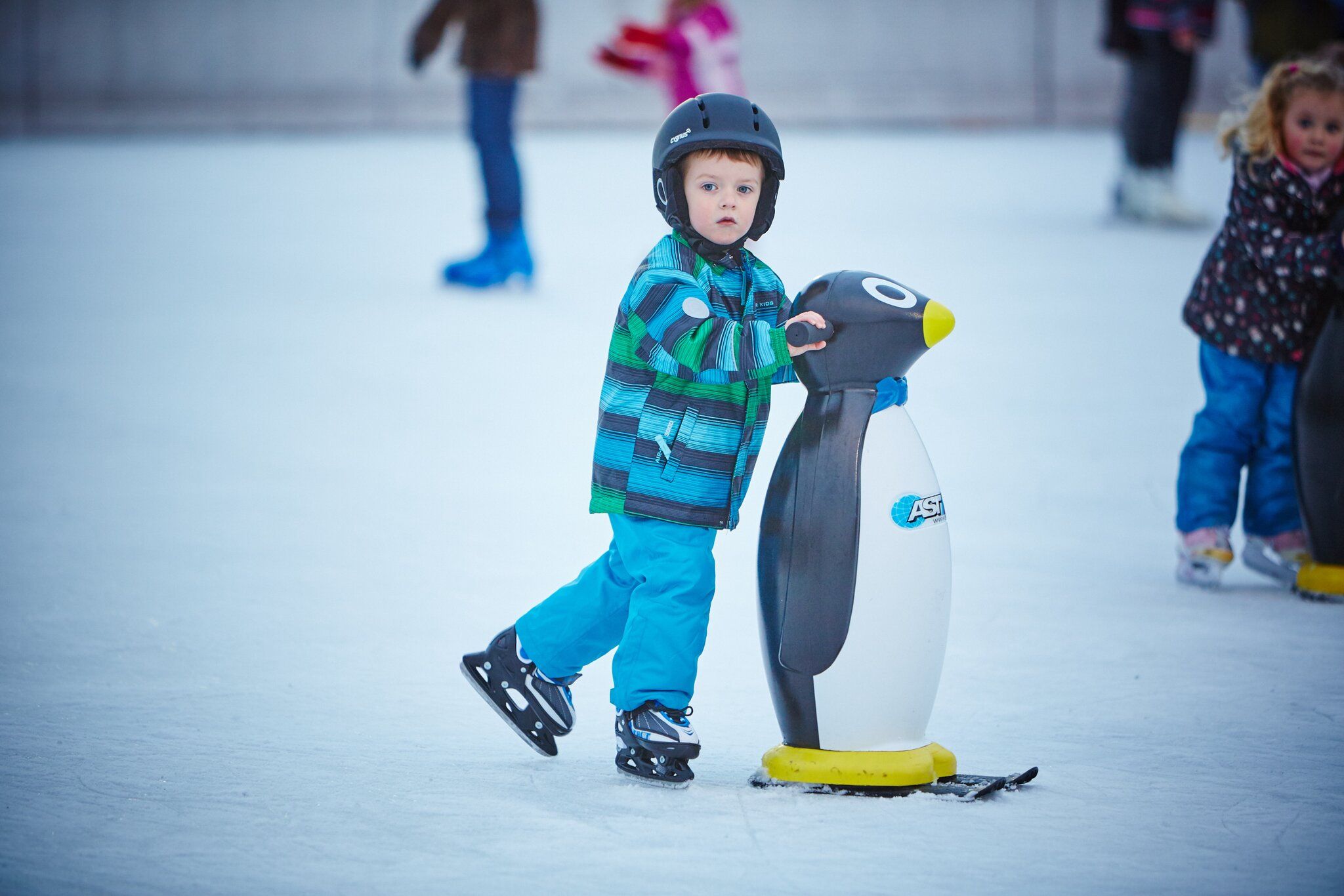A child wearing a helmet and colorful jacket holds on to a penguin skating aid on an ice rink.