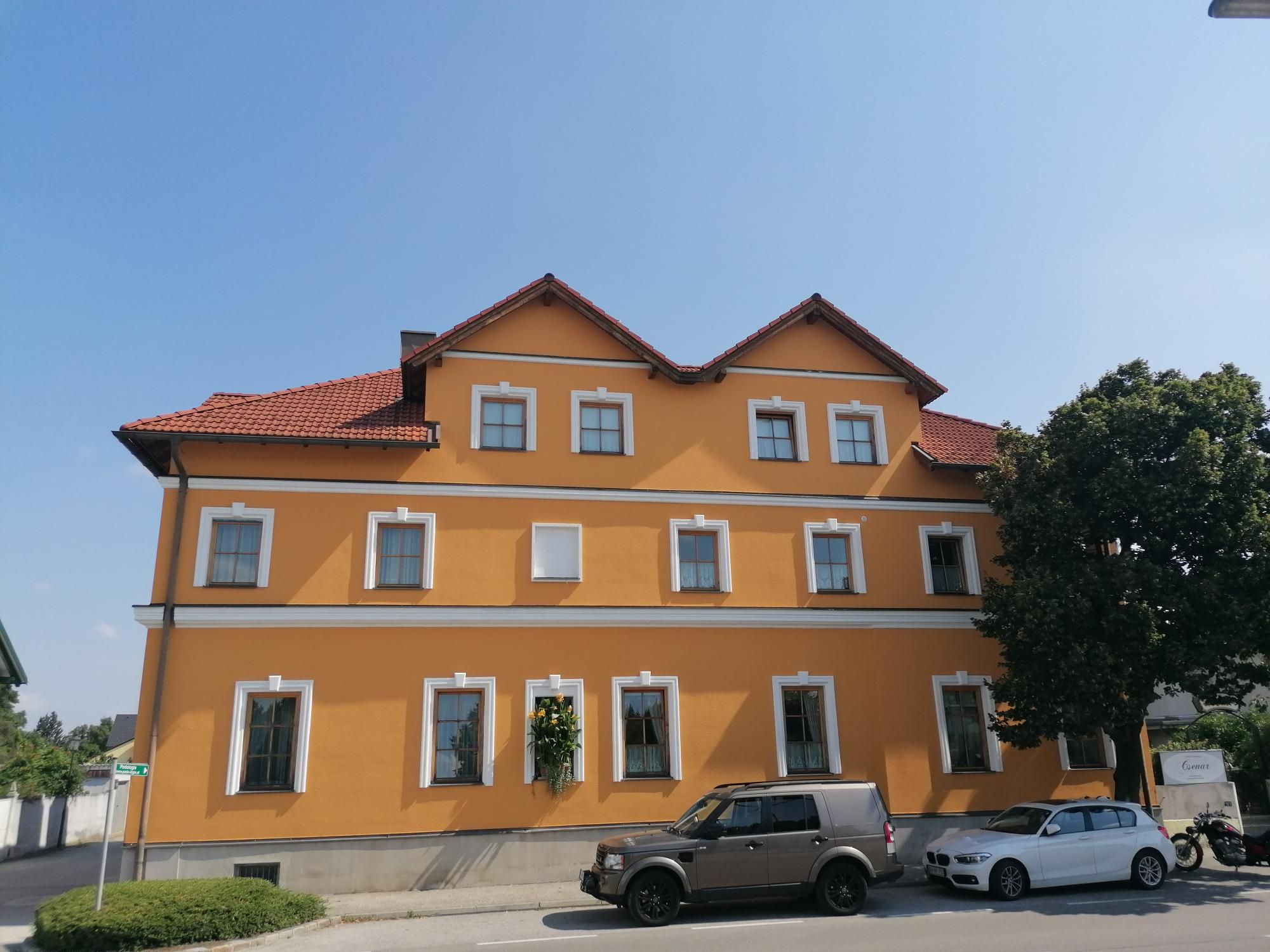 Yellow building with red roof tiles and several windows, cars parked in front of it and a tree.