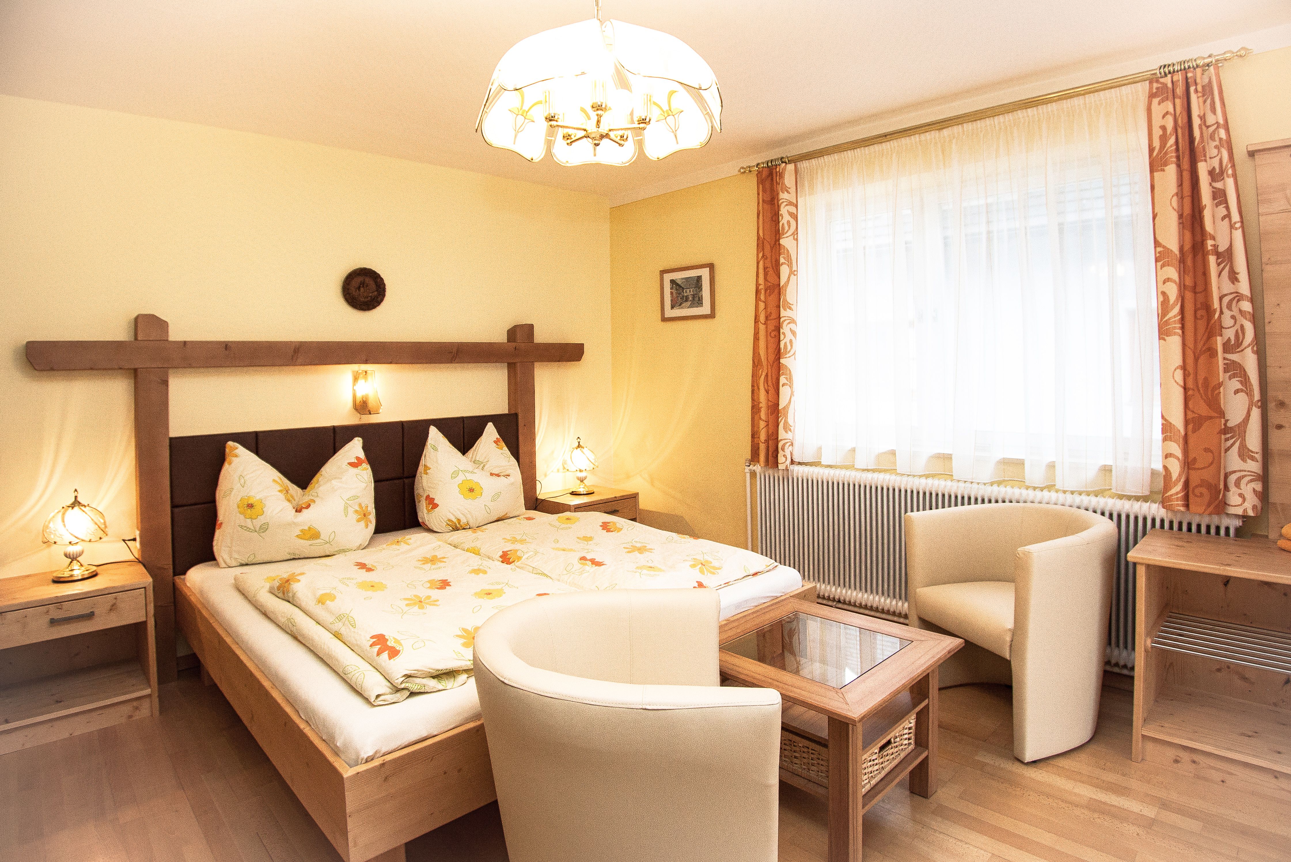 Cozy bedroom with double bed, two armchairs and a table. Bright curtains and warm lighting.