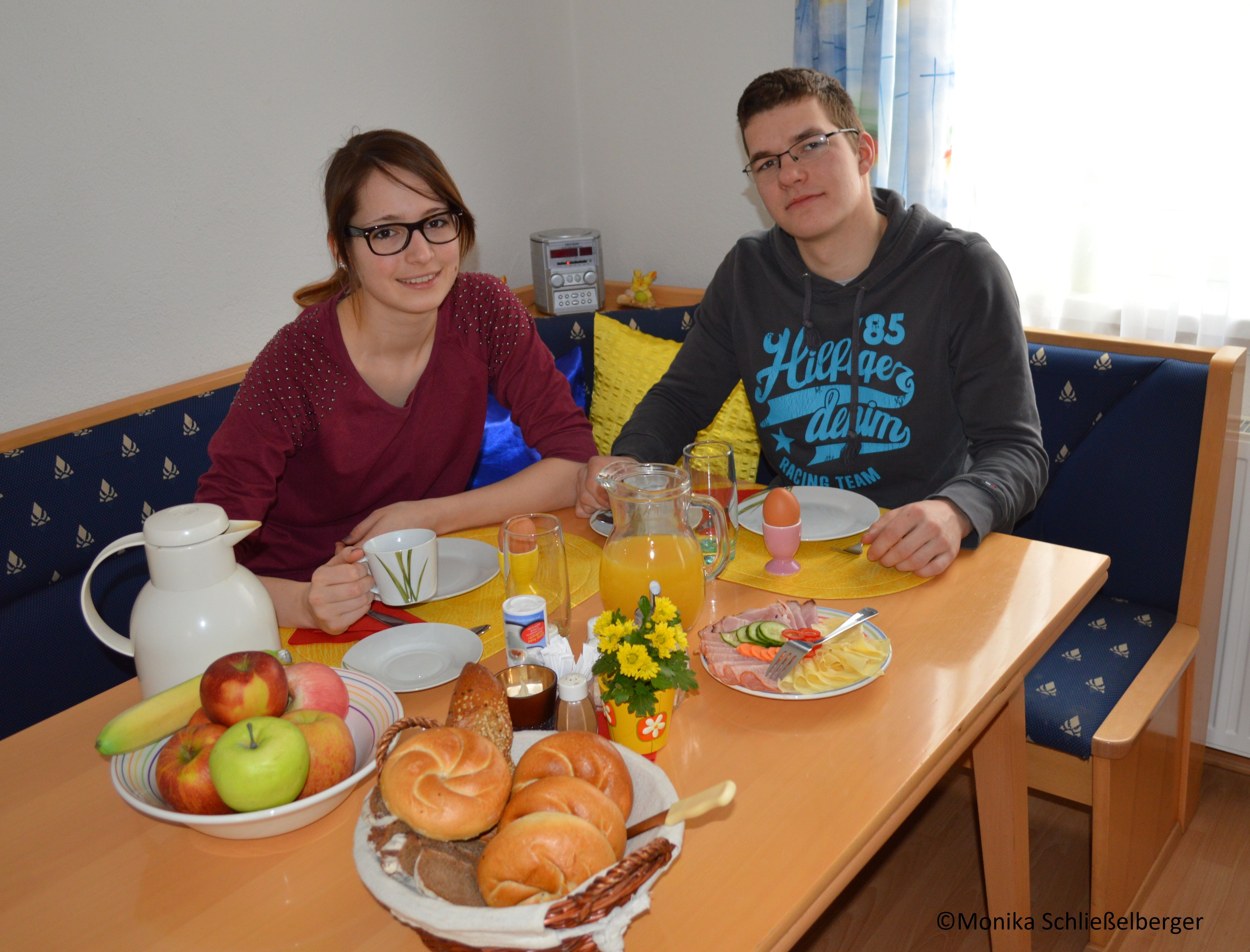 Two people sit at a table with breakfast, including bread rolls, fruit and juice.
