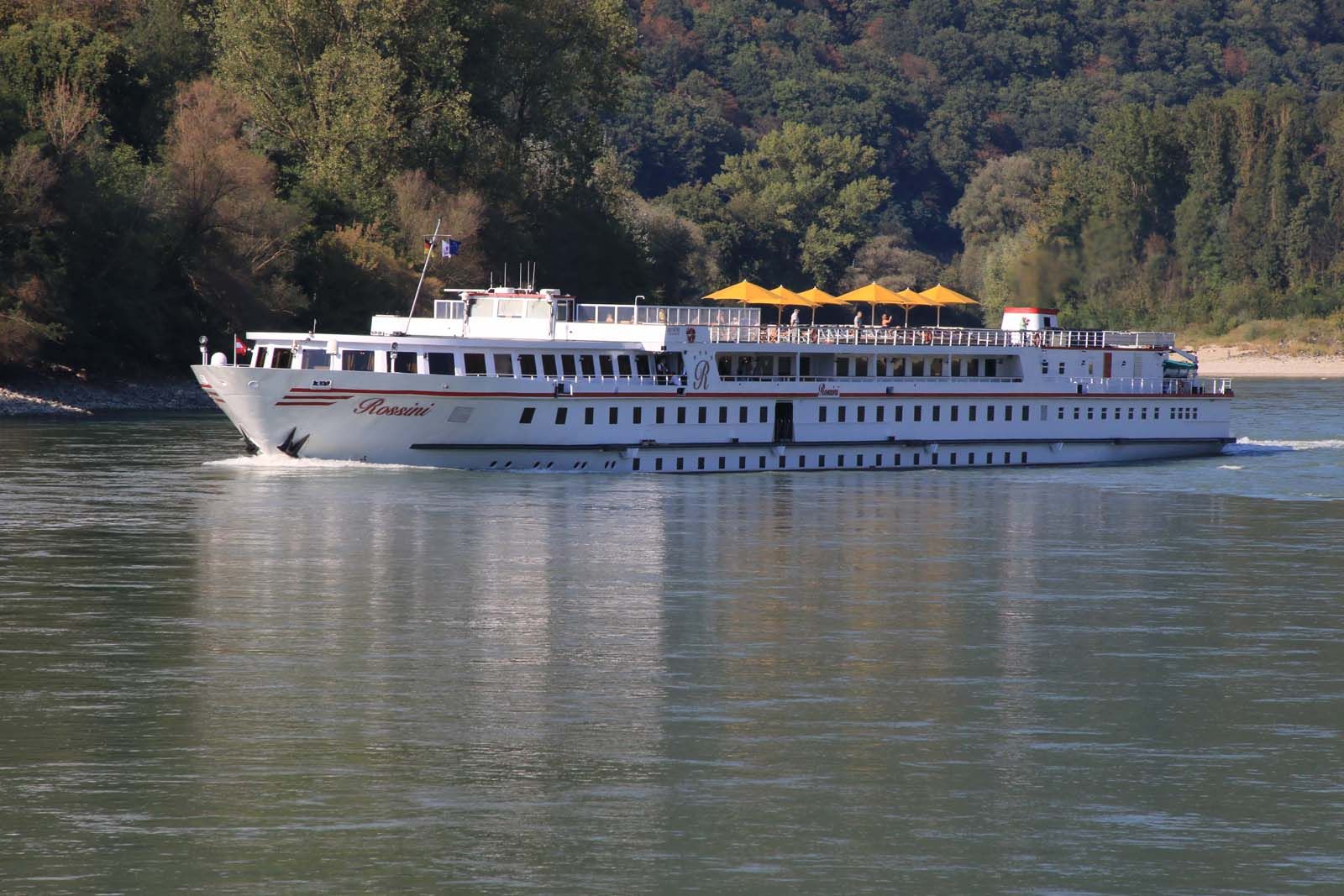 A river cruise ship named Rossini sails on the Danube, surrounded by wooded banks.