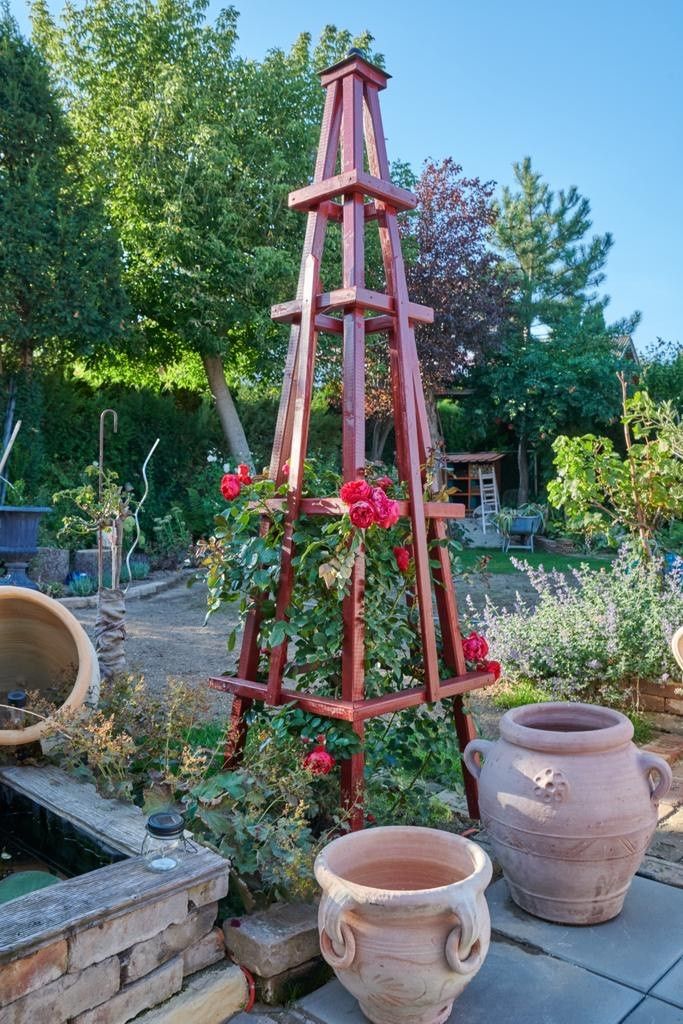 A garden with a red climbing support on which roses grow, surrounded by pots and plants.