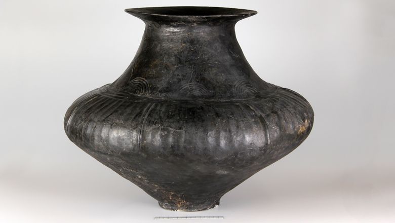 Large, dark conical-necked vessel with decorations on a light background.