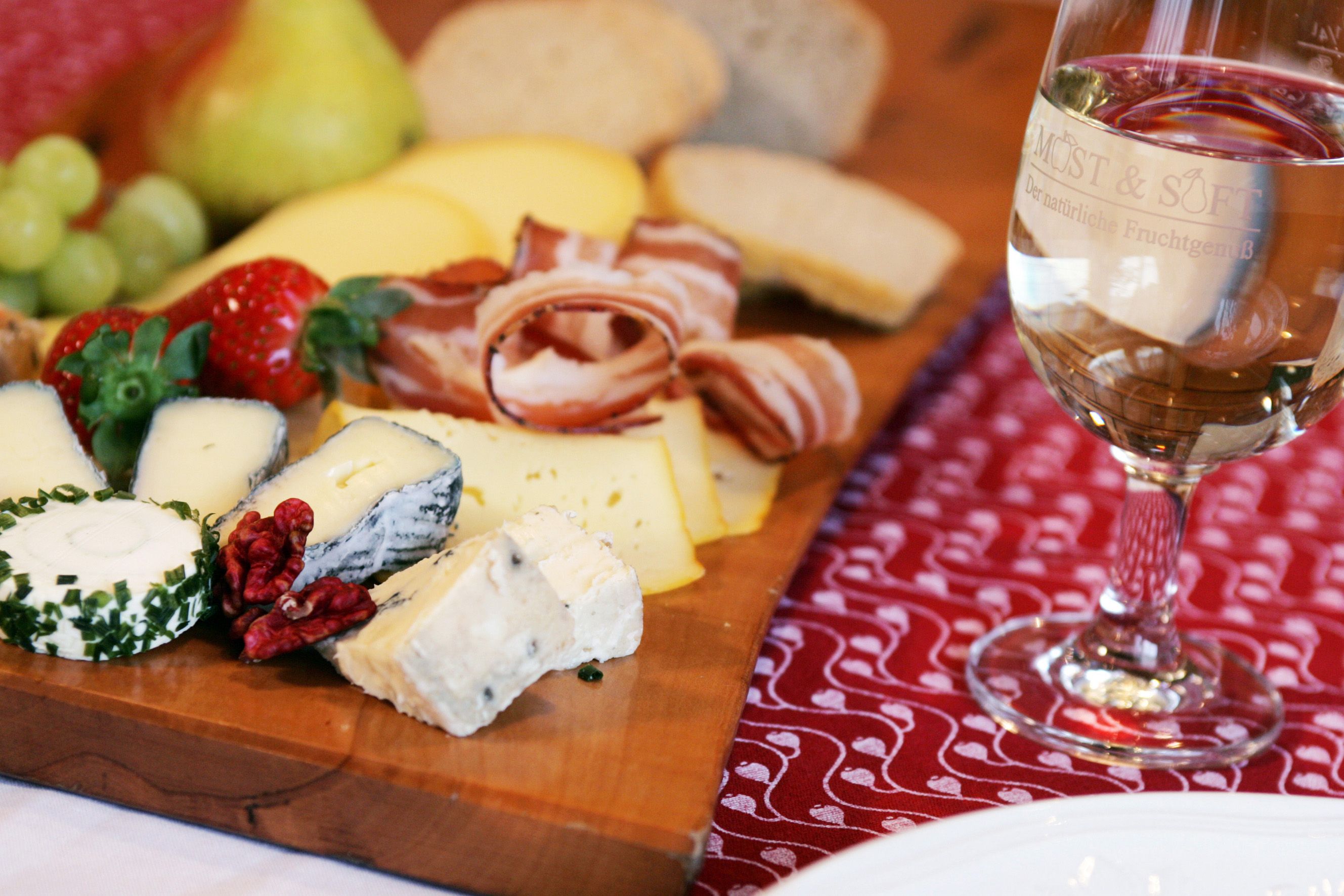 A snack with cheese, ham, bread and fruit on a wooden board, with a glass of white wine next to it.
